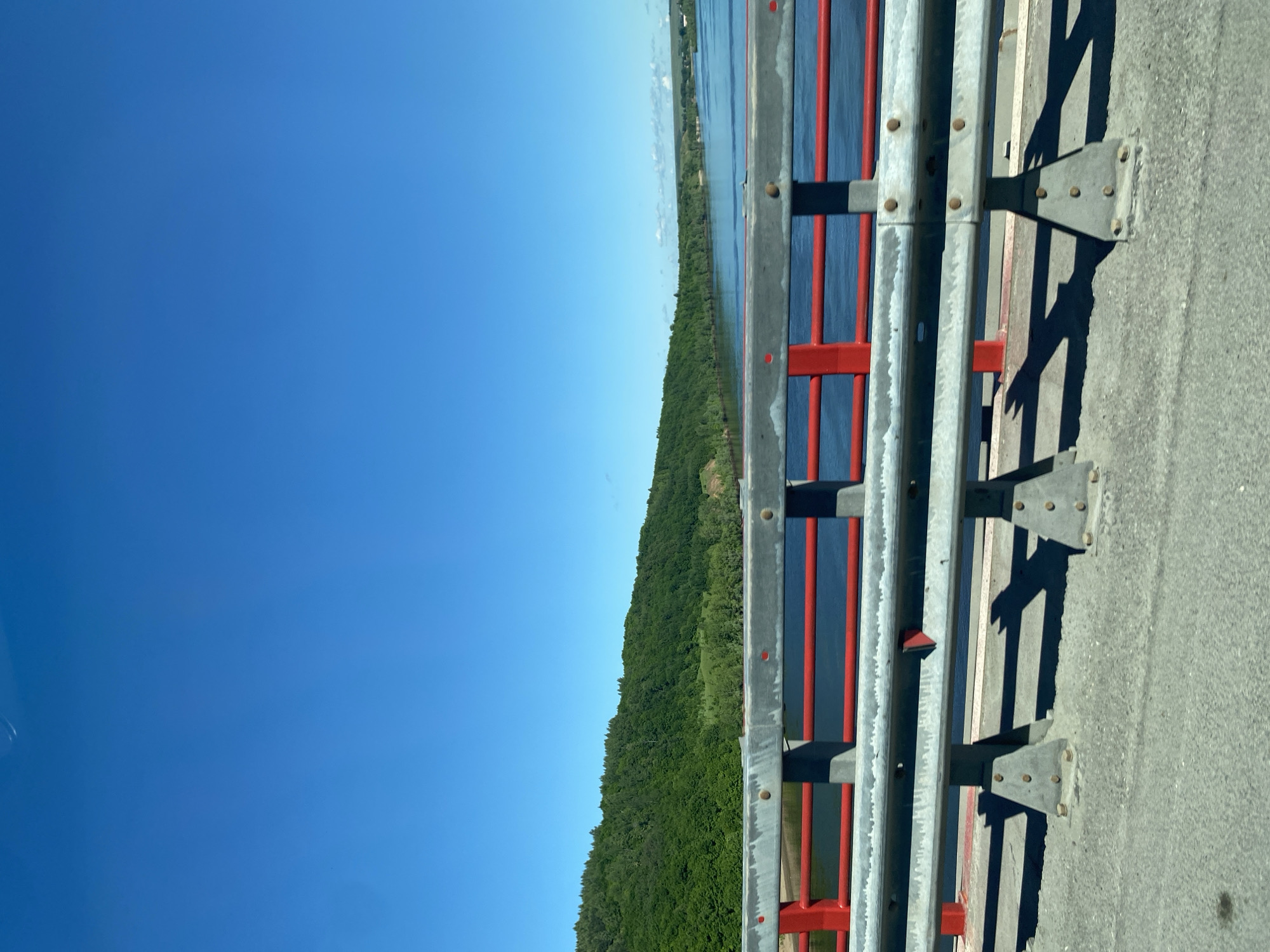 The image shows a roadside view from a bridge on a clear, sunny day, with the camera positioned looking along the bridge railing. The railing is gray with red vertical supports. Beyond the railing is a body of water and a heavily forested shoreline. The sky is a clear, bright blue. The location is likely near Sovkhoza "Pyatiletka", Russia, as requested.