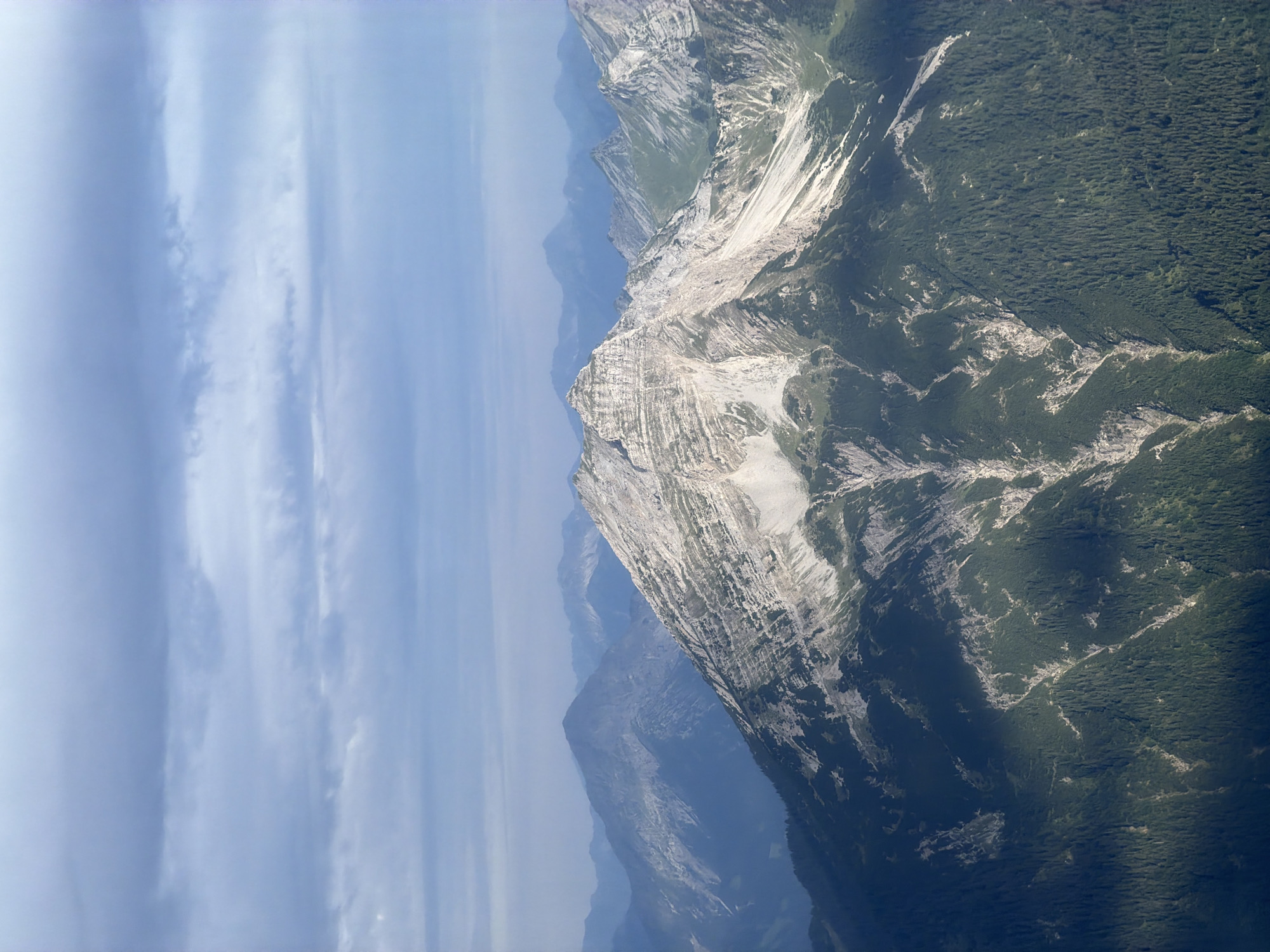 The image is an aerial view of a mountain range in Grainau, Germany, under a cloudy sky. The foreground is dominated by a steep, rocky mountain slope covered in dark green coniferous trees. Sunlight casts strong shadows across the lower slopes, highlighting ravines and rocky outcrops. The central mountain is a prominent peak with a jagged, rocky summit, showing layers of stone and patches of vegetation. In the background, several other mountain peaks fade into a hazy blue distance, with the sky above filled with horizontal streaks of grey and white clouds. There are no people or visible man-made structures in the image.