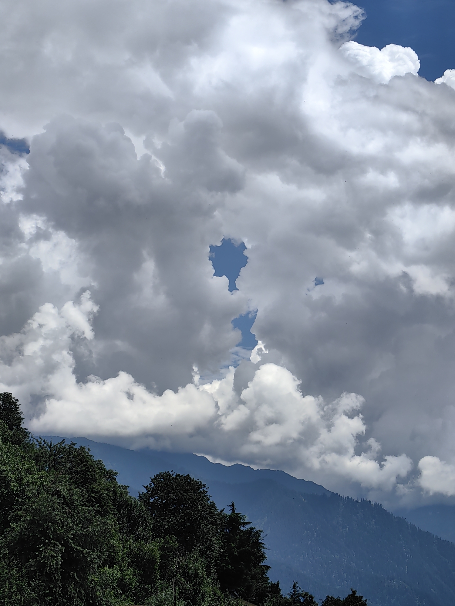The image depicts a scenic mountain landscape with a dramatic sky. In the foreground, lush green trees and foliage cover the lower left portion of the frame, leading towards a steep, tree-covered mountain slope. The middle ground features a hazy, blue-hued mountain range. The sky above is dominated by large, billowy clouds, with patches of blue sky visible. One striking feature is a distinct, irregular opening in the clouds, revealing a clear view of the blue sky beyond. The scene appears to be taken from a high vantage point, overlooking the mountainous terrain.