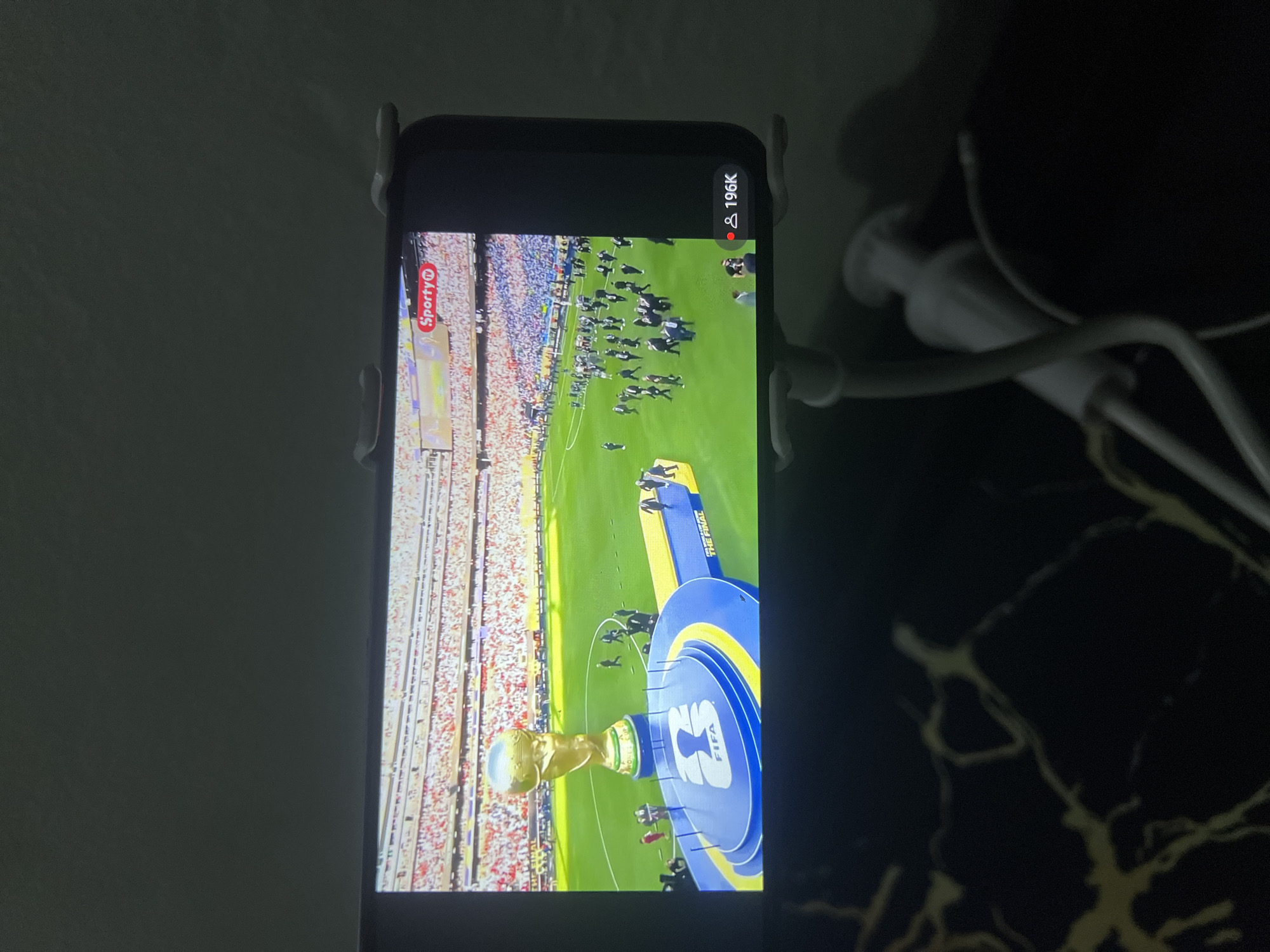 A mobile phone displays a scene of a soccer stadium. The screen shows the FIFA World Cup trophy on a podium in the center of the field. Spectators fill the stands, and people are walking on the grass. The word "Sporty" is visible in the upper right corner of the screen, suggesting a sports streaming or betting platform. Below the screen, a dark surface with yellow veining, likely a table, is visible. The background is dark, indicating low lighting conditions. The phone is held by a white stand. There is no indication of the city of Jos, Nigeria, in the image.