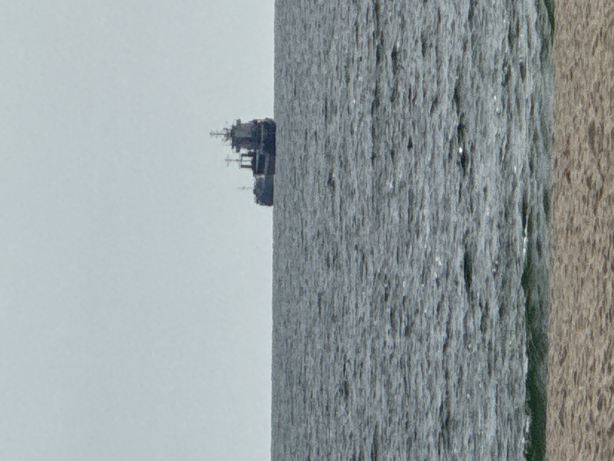 An eye-level, outdoor shot captures a marine scene, likely off the coast of Varna, Bulgaria, as suggested by the presence of a naval vessel. A significant portion of the frame is taken up by the sea, with its textured, grey-blue surface indicating moderate wave action. To the right, a strip of sandy beach is visible, its color a light tan. A warship or a large naval vessel is located off the coast. The sky is overcast, presenting a uniformly grey background that blends seamlessly with the sea's horizon, emphasizing the flatness of the scene. The elements combined portray a calm day.