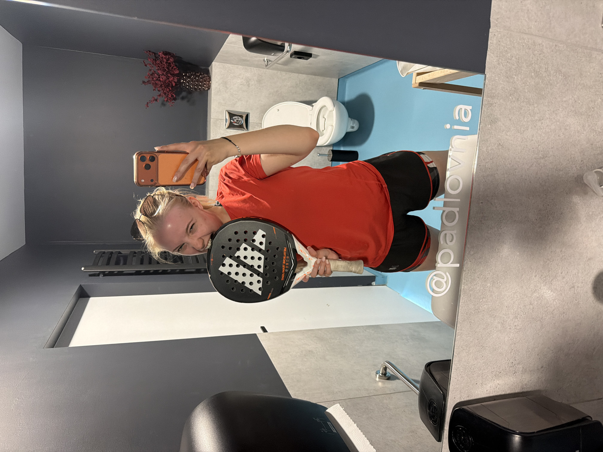 A person is taking a selfie in a bathroom mirror while holding a paddle tennis racket. The person has blond hair and is wearing sunglasses on top of their head, a red t-shirt, and black shorts with a red trim. They are holding a phone with an orange case in their right hand. The racket is black with white designs and is held in their left hand. The bathroom has gray walls, blue accents, and a toilet in the background. A sign on the mirror reads "@padlovnia". The location of the bathroom is Unknown, Unknown.
