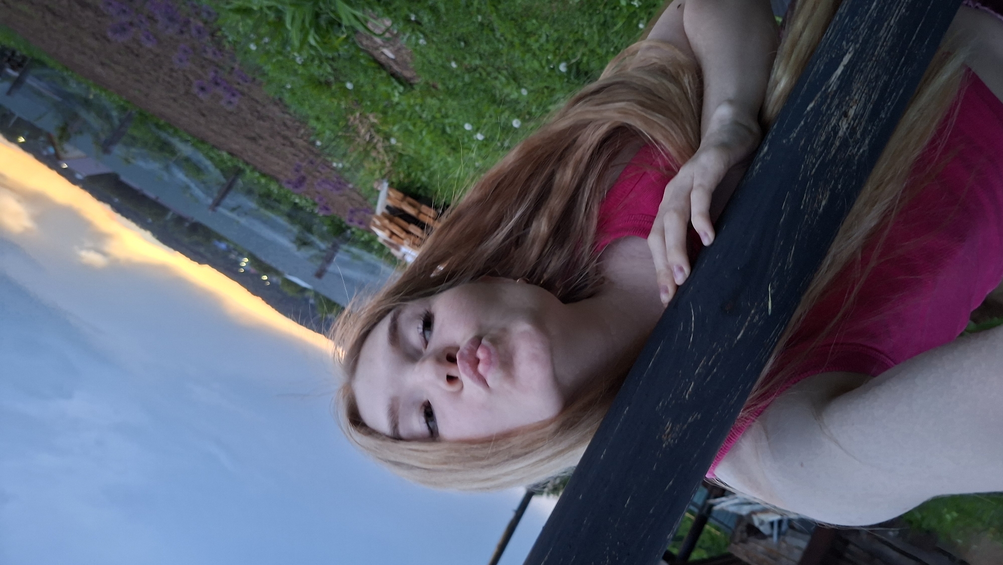 A young woman with long, light brown to blonde hair is captured in a close-up, outdoors during what appears to be either sunrise or sunset. She is wearing a bright pink sleeveless top and is positioned with her face tilted slightly upward, making a puckered or "kissing" expression towards the camera. Her right hand and arm are resting on a dark, rough-hewn wooden beam or fence post that diagonally traverses the image.
The background on the left features a sky transitioning from clear blue to a warm golden-orange, indicating the golden hour. A faint line of distant land or structures can be seen along the horizon. The upper right and background behind the woman reveal green grass interspersed with small white flowers, as well as clusters of purple flowers, possibly growing on or near another wooden structure. The overall setting is natural and open, suggesting a rural or semi-rural environment in Большой Букор, Russia. There is no visible text in the image.