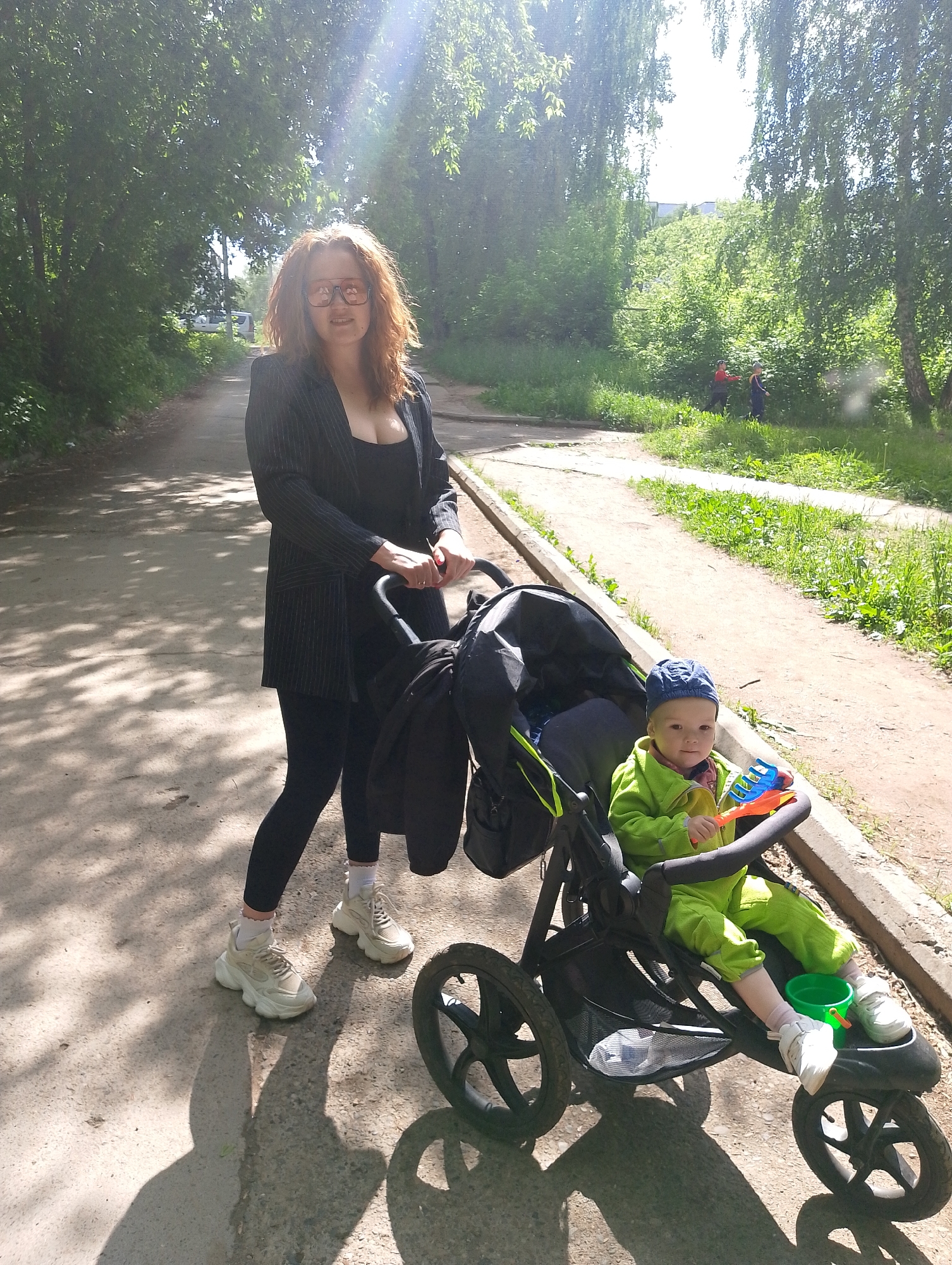 The image shows a woman standing on a paved path with a stroller in front of her. A young child sits in the stroller, wearing a blue cap and a green outfit, holding a toy shovel and a green bucket is visible near the child's feet. The woman is wearing a black top, black leggings, a black blazer, and light-colored sneakers. The path is shaded by trees on both sides. A grassy area is adjacent to the path on the right side, and there are two figures in the background in the grassy area. The location is in the city of Unknown, Unknown.