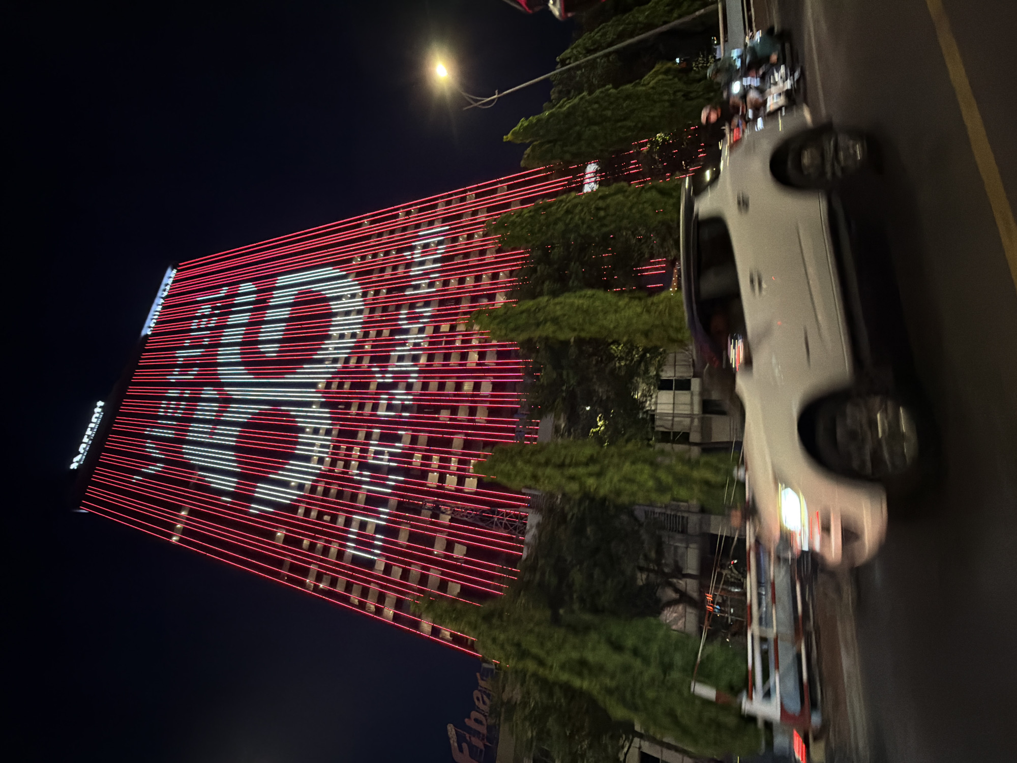 At night, a high-angle, medium shot reveals the Marriott Hotel in Đà Nẵng, Vietnam. The building’s façade is illuminated with red and white horizontal light stripes, creating a stylized representation of the Vietnamese flag. A white vehicle is prominently positioned on the right side of the frame, blurring due to motion. A street light shines above trees in the middle ground. Pedestrians are near the moving vehicle. In the lower-left corner, a partial view of a sign with "Fibers" is visible. The dark sky provides a contrast to the bright lights.