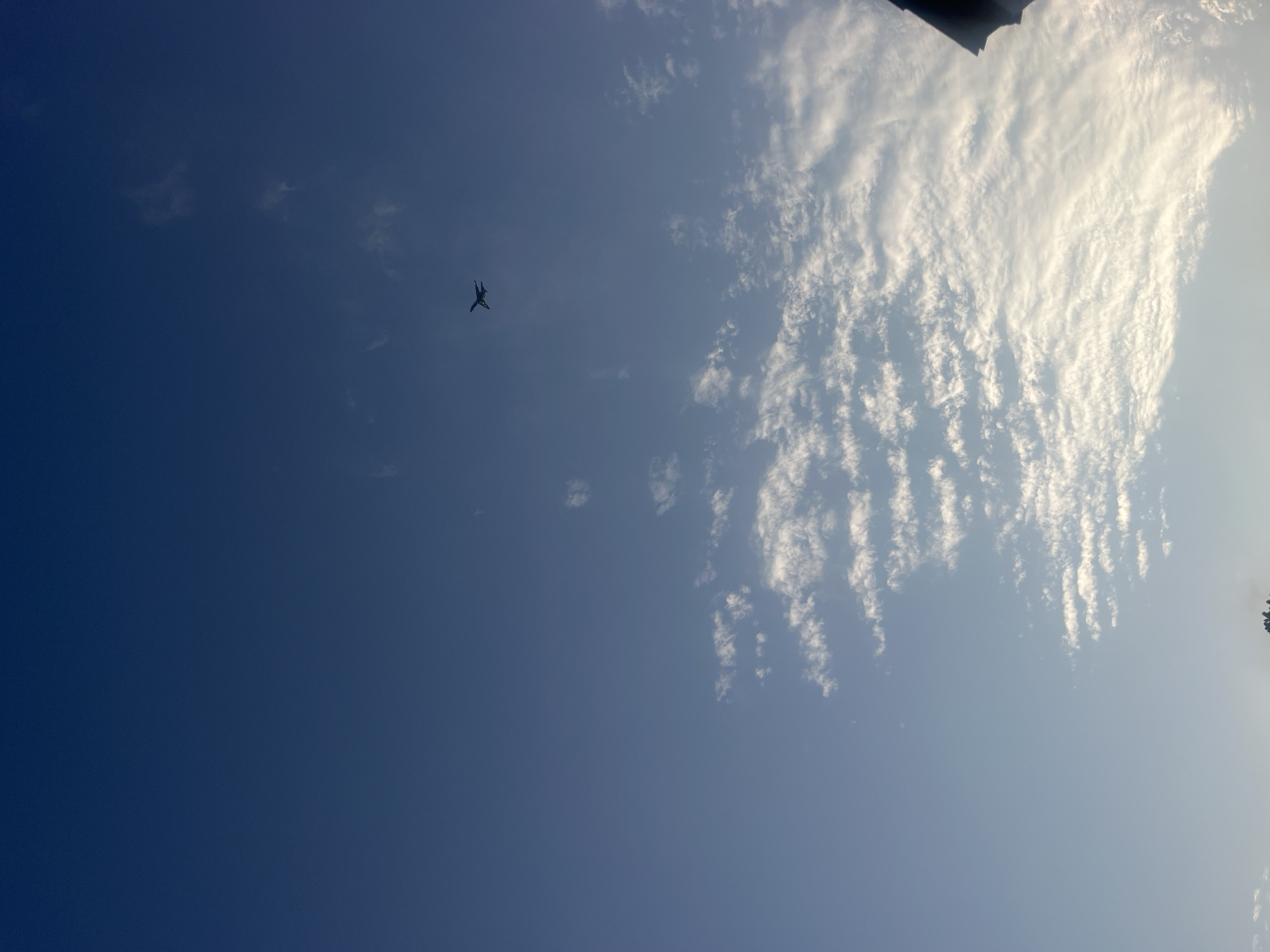 An airplane is visible in the sky. The sky is a gradient of deep blue at the top, transitioning to a lighter blue towards the bottom. There are scattered white clouds. A dark object, possibly a rooftop or an awning, is visible in the lower right corner of the image. The image appears to be taken outdoors in Jacksonville, United States of America.