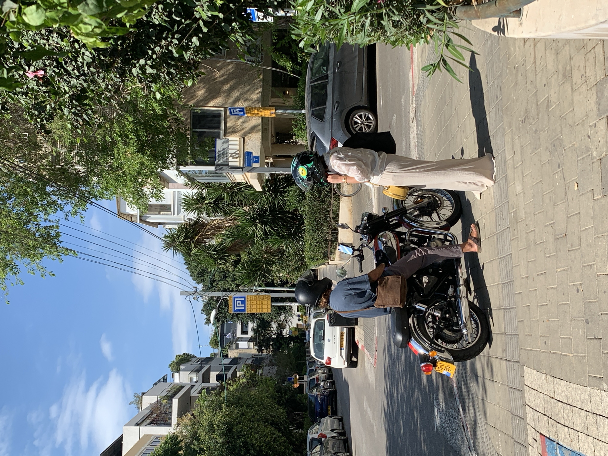 Two people are standing with two motorcycles on a sidewalk next to a street in Tel-Aviv, Israel. The street is lined with parked cars. The motorcycles are parked near a curb. The people are dressed in casual clothing and one of them is wearing a helmet. There are buildings and trees in the background. A parking sign is visible.
