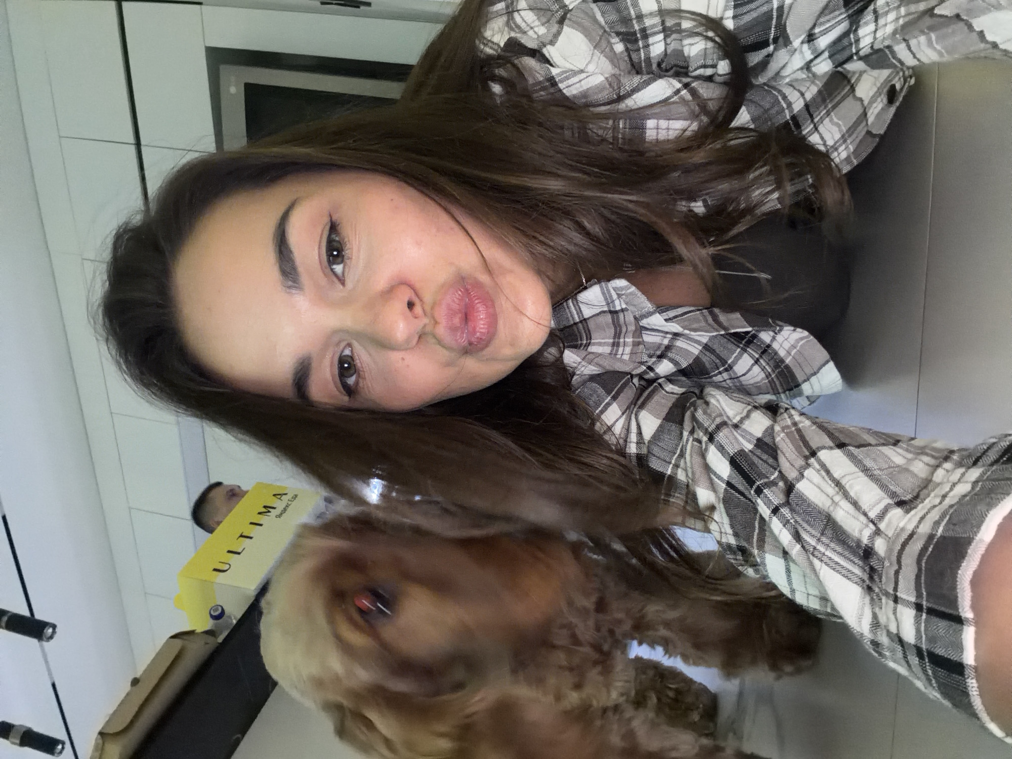 A woman with long brown hair and a plaid shirt poses for a selfie with a brown dog. She is pouting her lips. The dog is in the lower left corner of the frame. Behind the dog and woman is a yellow box with the word "ULTIMA" printed on it. In the background are white cabinets and countertops, suggesting the location is likely a kitchen. Based on the Cyrillic text "Яндекс Еда" on the box, it is possible the location is in Russia, perhaps Krasnoyarsk. A man's head is also visible behind the box.