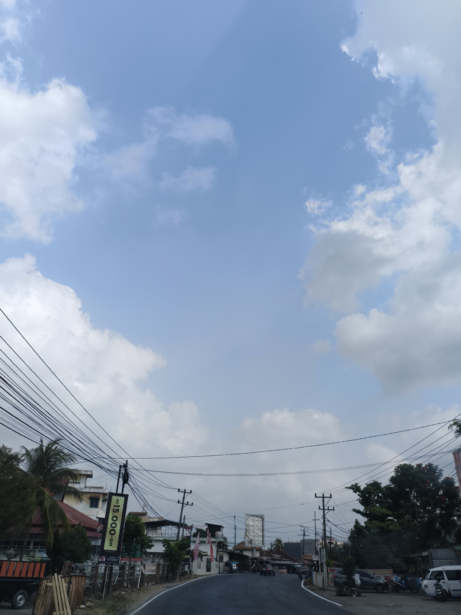 This image captures a street scene in Kemala Raja, Indonesia, under a partly cloudy blue sky. The foreground features a paved road curving to the right. On the left side of the road, a billboard displaying "Rp15.000" and "on" is prominently visible, along with a cluster of utility poles and a dense network of overhead wires. A palm tree and some residential or commercial buildings with red roofs are also situated on the left. A truck with an orange flatbed is parked on the left shoulder. On the right side of the road, more utility poles and wires are present, along with lush green trees and several parked vehicles, including a white van. The overall impression is of a somewhat cluttered, but functional, urban environment.