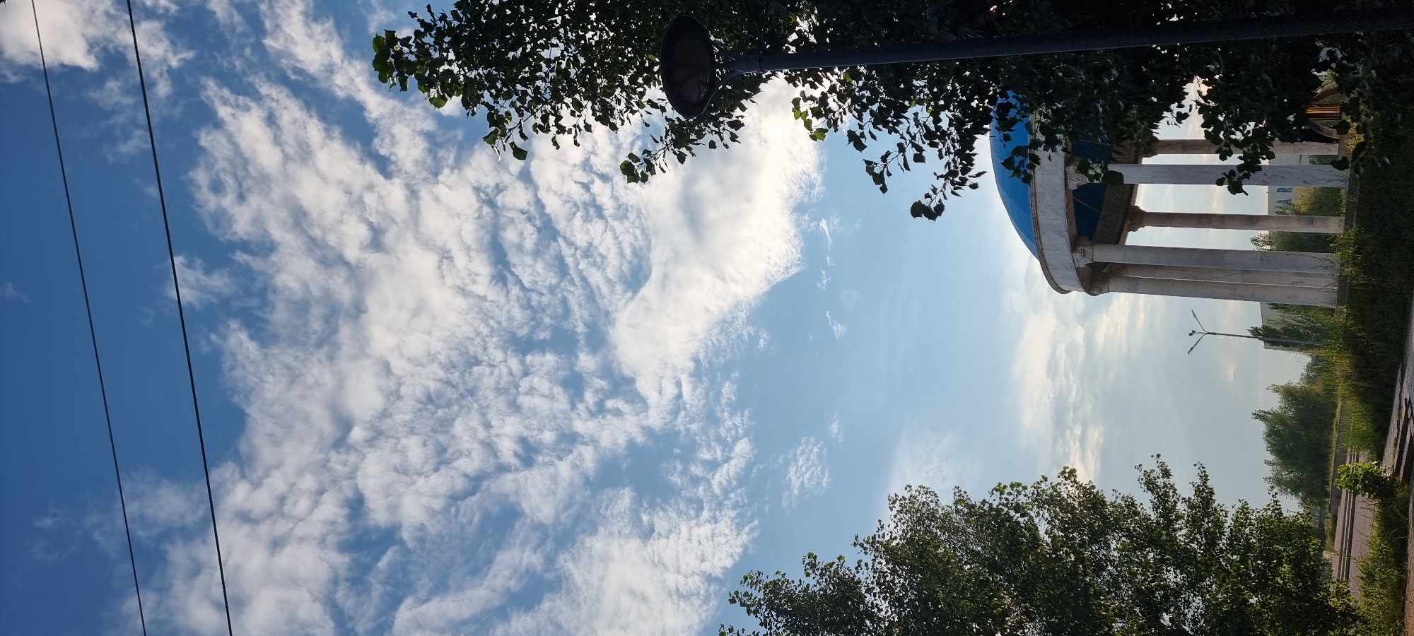 The image shows a bright blue sky filled with scattered white clouds, with two dark power lines crossing horizontally across the upper portion of the frame. In the lower right, a tall, dark lamppost is partially obscured by the dense green foliage of a tree. To the left of the lamppost, a white structure with a bright blue dome roof is visible, supported by several tall, fluted white columns. This structure appears to be an open-air pavilion or gazebo. In the background, there are more trees and some low-lying buildings. The ground in the foreground is covered with green grass and some paved pathways. This scene is likely located in Aktobe, Kazakhstan, possibly in a park or public space given the presence of the pavilion and lamppost. There are no people visible in the image.