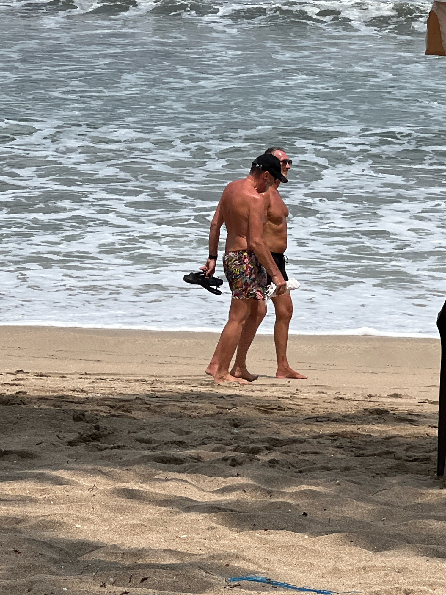 In Kuta, Indonesia, two shirtless men walk along the sandy shore. The man on the left wears patterned swim trunks and carries a black sandal in his right hand. The man on the right, wearing black swim trunks, holds a white cloth in his left hand and a black baseball cap on his head. The ocean with its foamy waves extends behind them, meeting the beach at the shoreline. The foreground is dominated by the textured sand, casting shadows and showing debris.