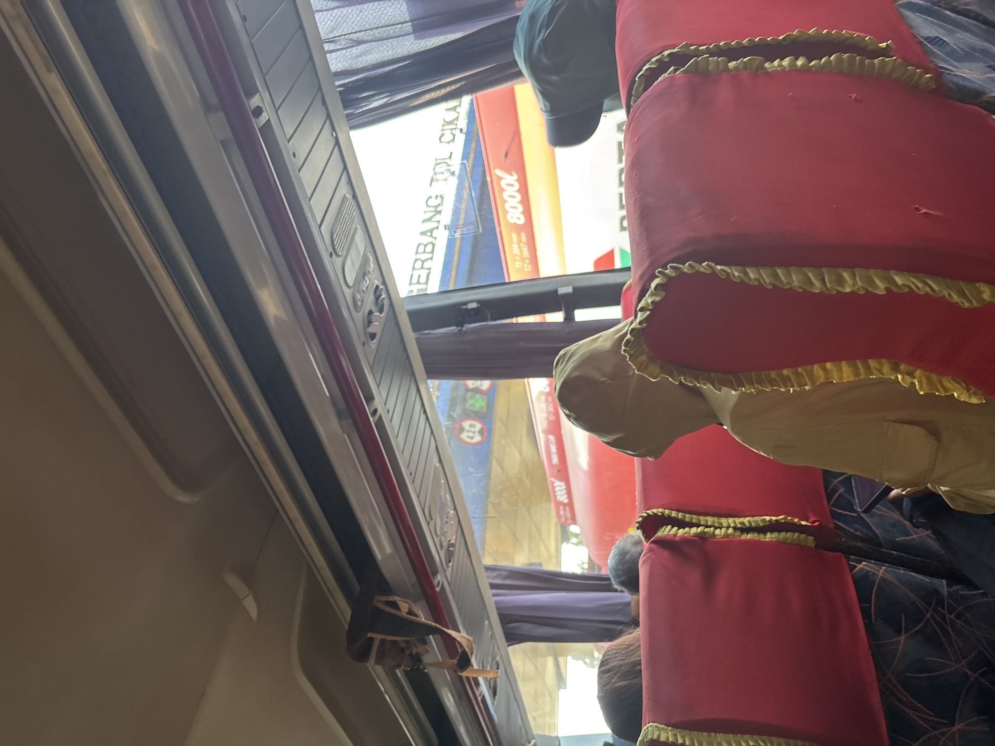 The image depicts the interior of a bus, with passengers seated. The seats are covered in red fabric and trimmed with gold decorative fringe. The overhead compartment is visible, as is a bag hanging from it. Through the window, another bus or vehicle is visible, with text that appears to be "GERBANG TOL CIKAMPEK" and "8000L" printed on its side. This suggests the bus is on a toll road in Indonesia, specifically near Cikampek, which is a major toll road interchange and town in West Java, Indonesia, not Purwakarta. The scene is taken from the perspective of a passenger seated towards the rear of the bus.