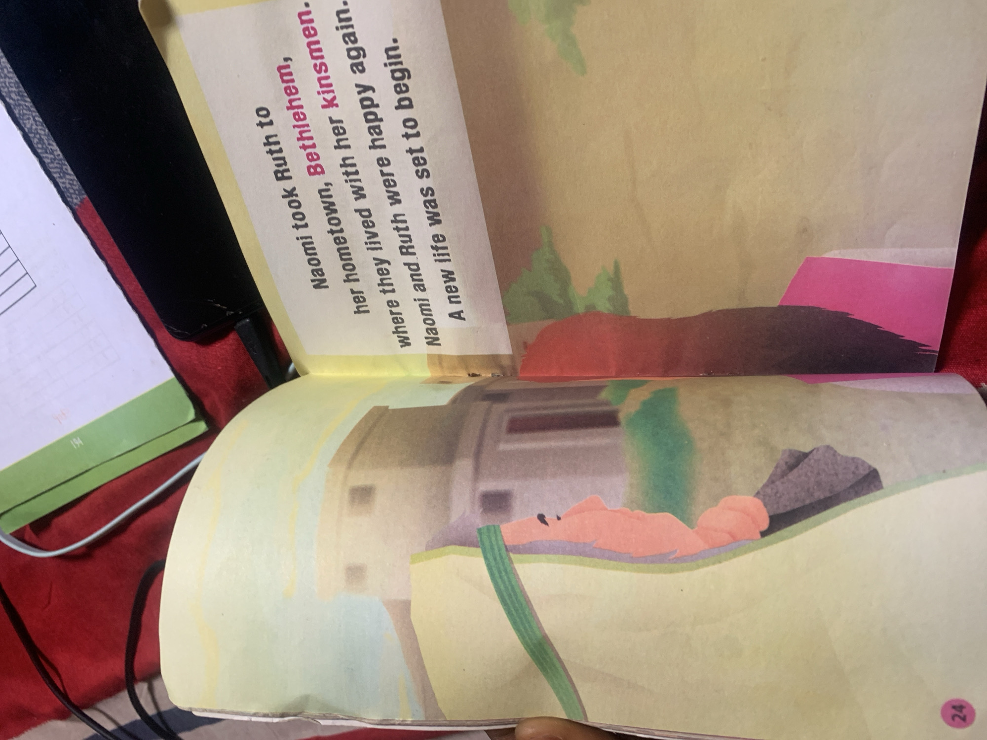An open children's book displays a scene depicting a story about Naomi and Ruth. The left page has an illustration of a woman in profile wearing a headscarf and a beige garment, with a building in the background. The number '24' is in a pink circle at the bottom left. The right page contains text that reads: "Naomi took Ruth to her hometown, Bethlehem, where they lived with her kinsmen. Naomi and Ruth were happy again. A new life was set to begin." The text is positioned over a plain background with subtle texture. The book is open on a red surface, with parts of a green folder and black cords visible in the upper portion of the image.