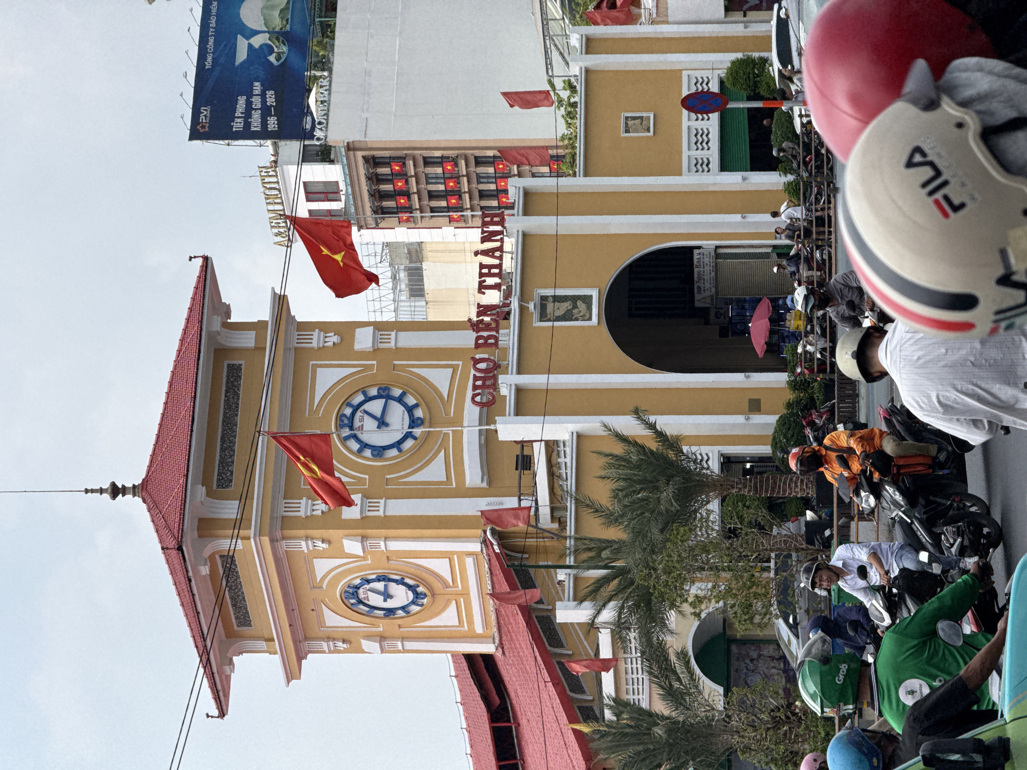 This image depicts the clock tower of Bến Thành Market (Chợ Bến Thành) in Ho Chi Minh City, Vietnam. The tower is predominantly yellow with white accents and a red roof, displaying two clock faces with blue markings. Vietnamese flags are prominently displayed on and around the tower. In front of the market, there are palm trees and a bustling street scene. People are seen riding motorbikes. The foreground is somewhat obscured by helmets and parts of motorbikes, indicating the perspective of someone on the street. Above the market, signs and advertisements are visible on adjacent buildings, including a large billboard with the text "TIÊN PHONG KHÔNG GIỚI HẠN 1996-2026."