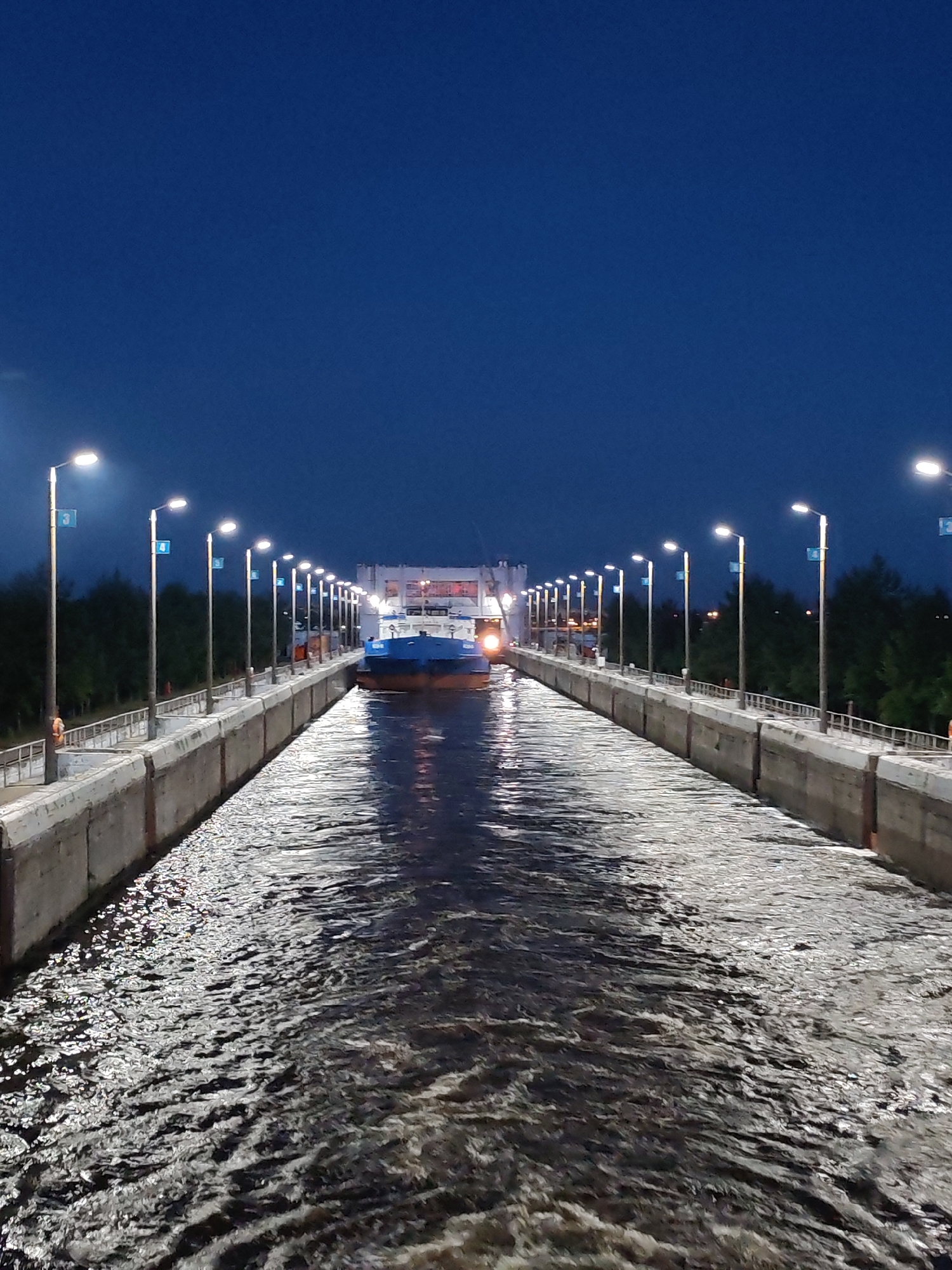The image depicts a vertical shot of the Sheksna river lock in Sheksna, Russia, at twilight. A large, blue and white cargo ship is centrally positioned within the concrete lock. Both sides of the lock are lined with concrete walls and numerous lampposts, each casting light onto the water. The sky above is a deep, clear blue, indicating nighttime or very late dusk. The water in the lock is turbulent, with ripples and reflections of the lights. Trees are visible in the background on the right side of the image.