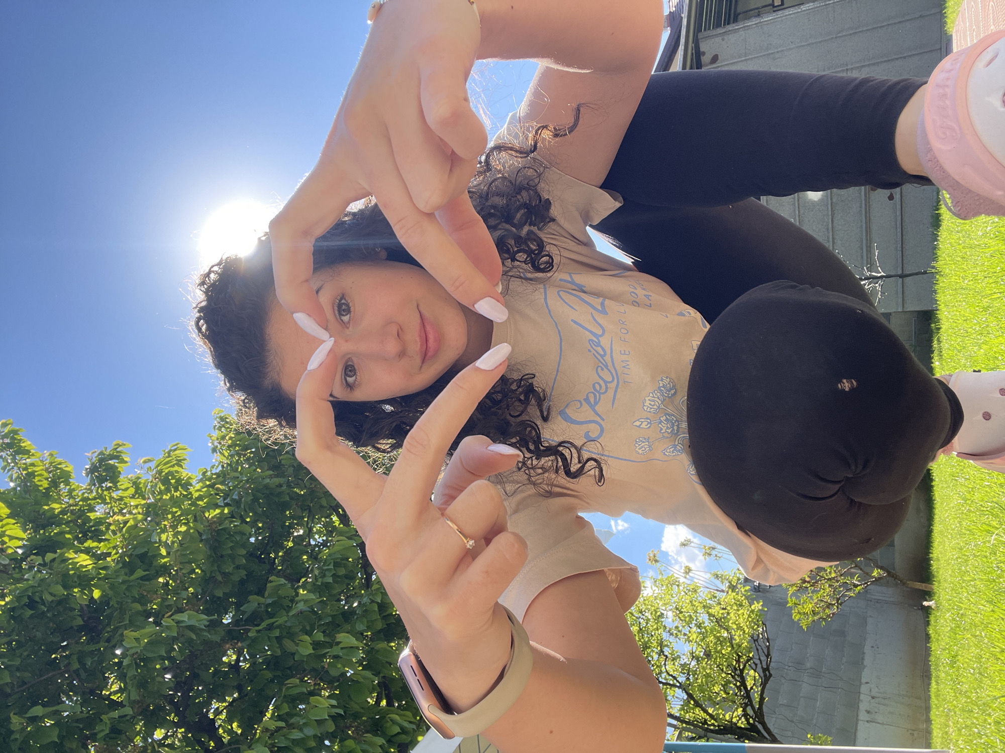A young woman with curly hair is taking a selfie outdoors. She is wearing a beige t-shirt with blue graphics, black leggings, and pink shoes. She is making a hand gesture with both hands in front of her face. She is wearing a ring on her left hand and a watch on her left wrist. The sky is bright blue with the sun shining. There are trees in the background. The location is believed to be in Sofia, Bulgaria.