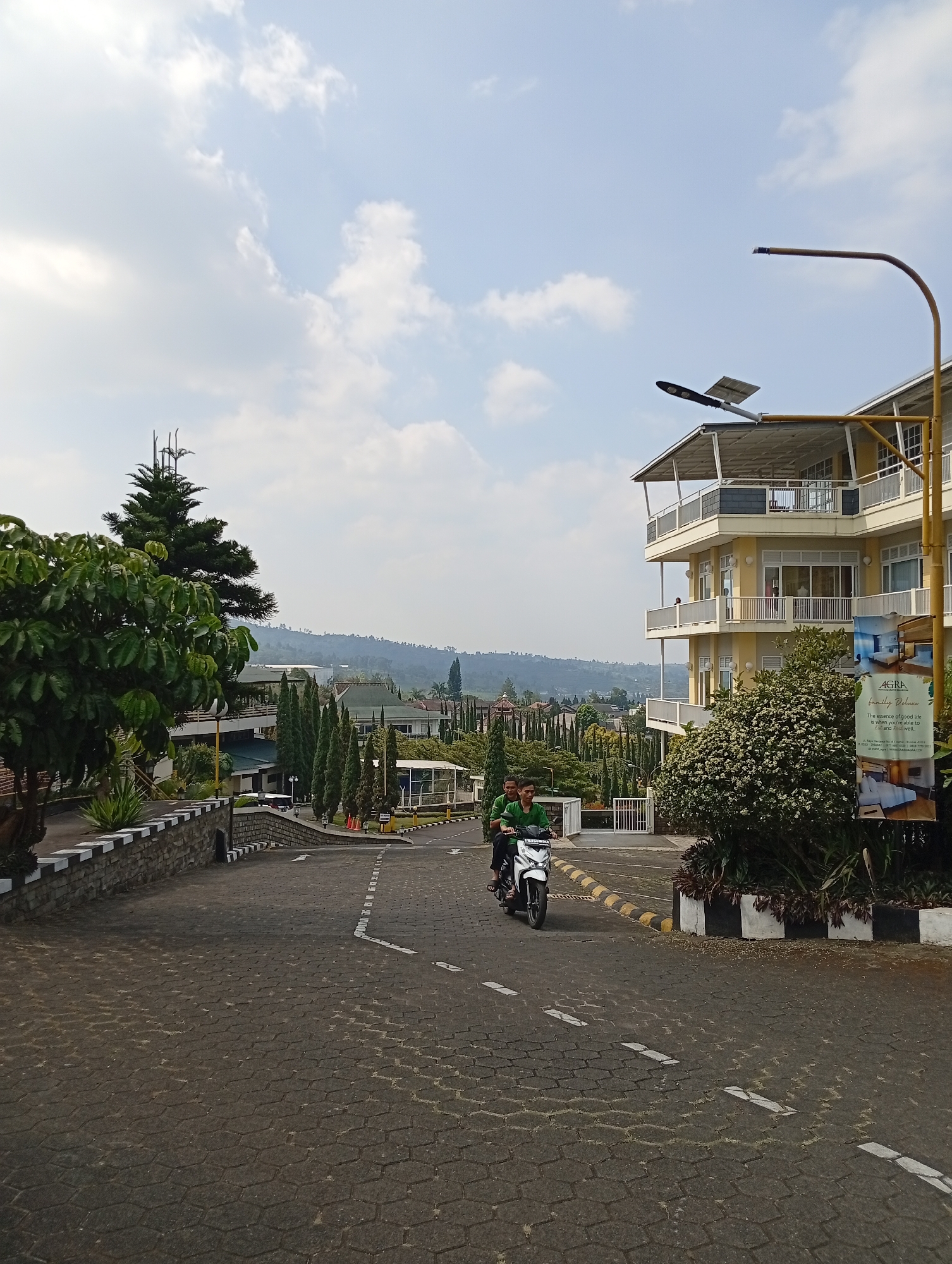 The image captures a paved road leading towards a multi-story yellow building with balconies. Two individuals are riding a white motorcycle on the road, with the passenger in front wearing a green shirt and the driver behind also in a green shirt. The road is made of hexagonal pavers and has white dashed line markings. To the left of the road, there's a stone wall with white and black trim and some greenery. A tall evergreen tree is visible behind the wall. The building appears to be a hotel or resort, indicated by the signage on the right side of the image which reads "AGRA family Deluxe". In the background, rolling hills covered in trees are visible under a partly cloudy sky. The scene is set in Cimacan, Indonesia.
