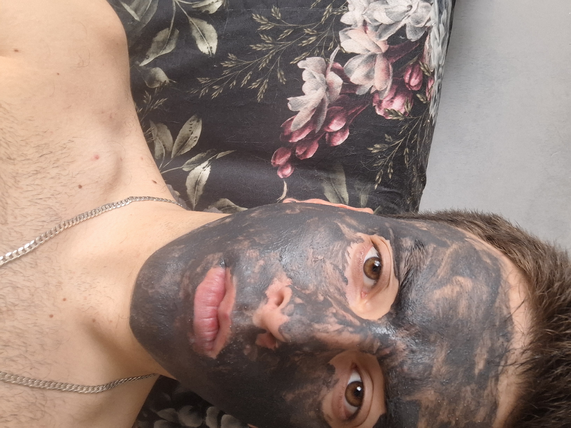 A human male with a black facial mask is lying on a pillow with a floral pattern. He is wearing a silver chain around his neck. The location is unknown, and there is no evidence it is in Sofia, Bulgaria.