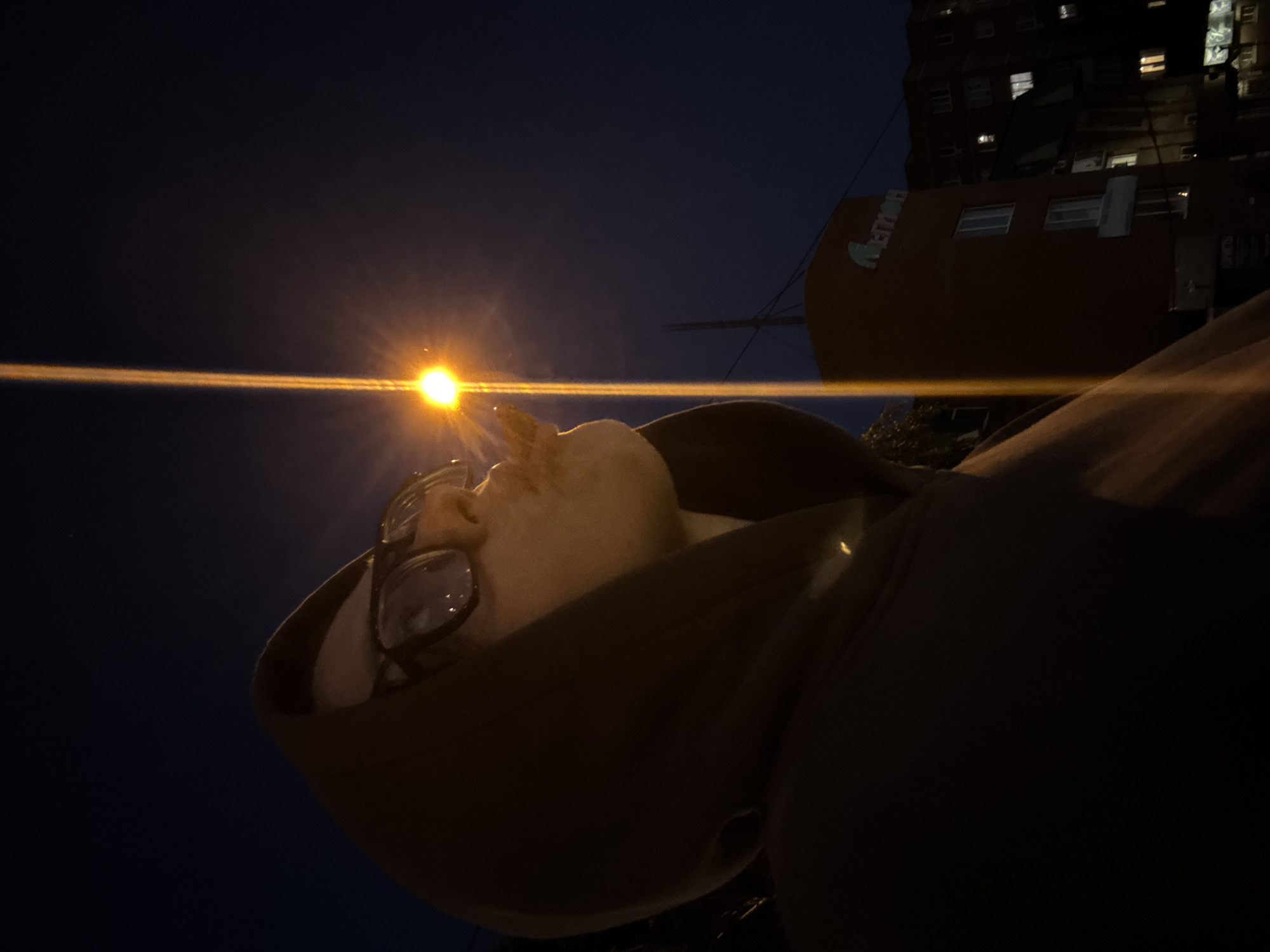 An outdoor, low-angle, night-time photograph depicts an individual in profile, facing right, from the chest up, in the city of Unknown, Unknown. The person wears a dark hooded sweatshirt with the hood up and clear-rimmed glasses, reflecting light from a prominent street lamp. Their lips are pursed, and a plume of white smoke or vapor is visible emanating from their mouth, illuminated by the light. A bright, yellow-orange street lamp with a significant lens flare creating a vertical light streak dominates the right half of the image, positioned centrally in the upper right quadrant. In the background on the right, a multi-story, reddish-brown building with numerous windows is visible. A sign with the Cyrillic word "АЛТЫН" (Altyn) is affixed to the upper portion of this building. Further behind and to the right, another building with lit windows is partially visible in the darkness. The sky is dark, indicating night.