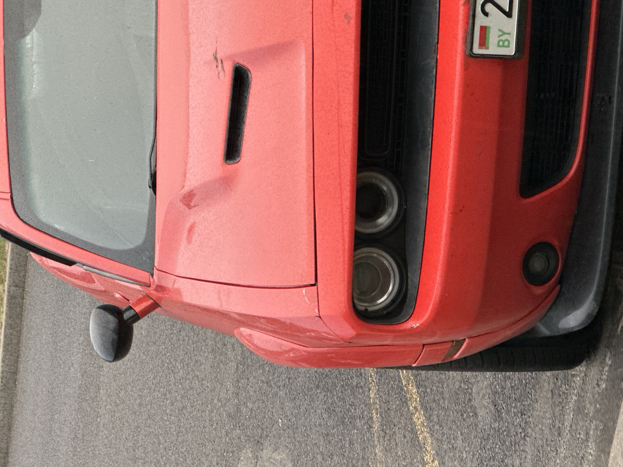 A red, widebody Dodge Challenger is prominently positioned, angled slightly towards the viewer, on an asphalt surface in Бараўлянскі сельскі Савет, Belarus. The vehicle's front passenger side is visible, displaying a dusty red paint finish with minor scuffs and dirt accumulation on the hood and fender. A black, oval-shaped side mirror extends from the passenger side A-pillar. The windshield is visibly dirty. The hood features a black, rectangular air intake scoop. Below the hood, dual circular projector headlights are recessed into black housings. The front bumper is red, with a circular fog light on the passenger side. A black lower grille and a dark grey front splitter are also visible. A portion of the front passenger tire is seen. A white Belarusian license plate, partially visible on the lower right of the front bumper, displays the Belarusian flag, "BY" in green text, and the numeral "2". The asphalt ground exhibits faint yellow painted lines and appears textured or wet. A concrete curb runs along the top left edge of the frame, bordering the asphalt. No individuals are present in the scene.