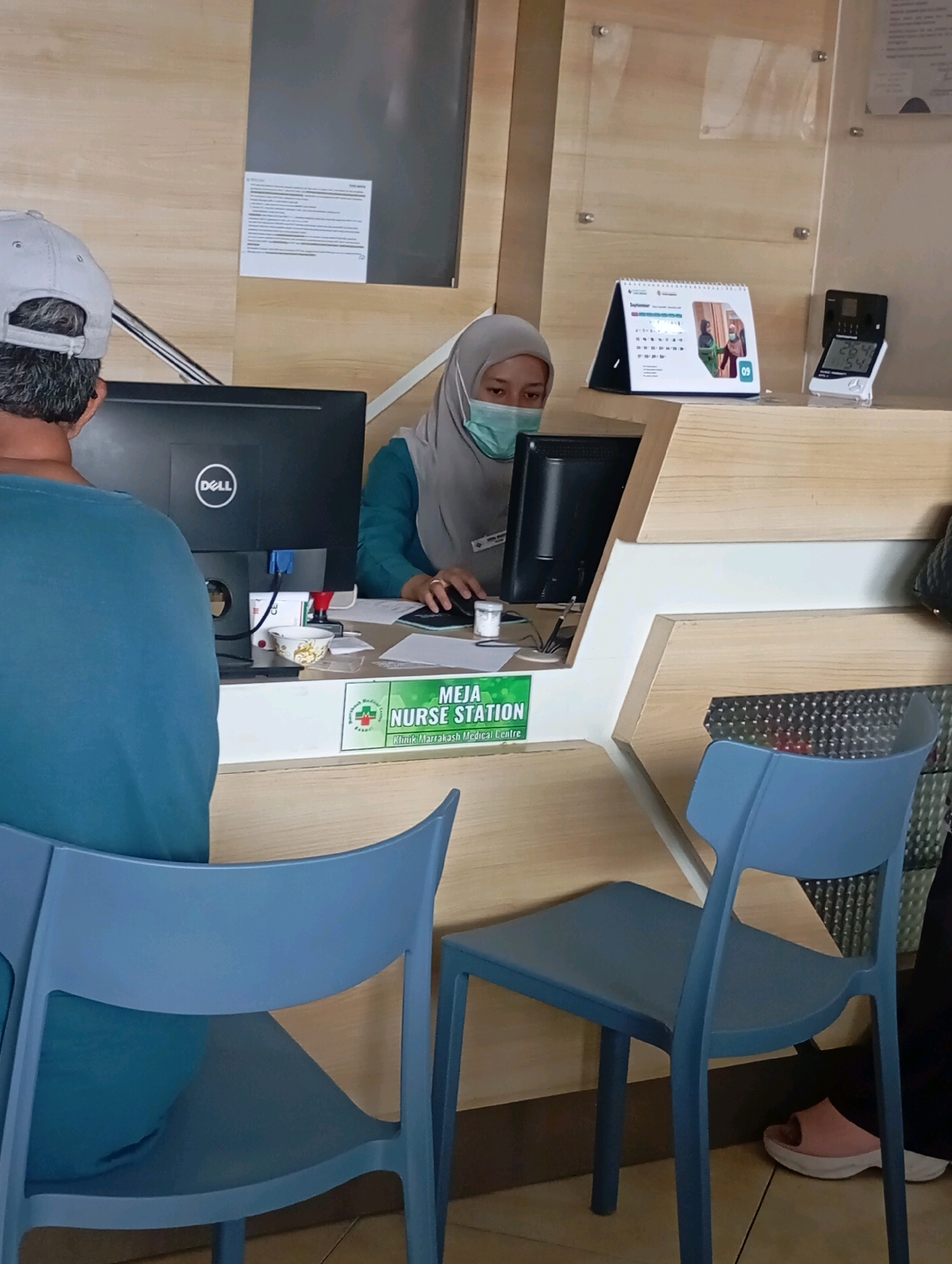 The image shows a nurse station at a medical center. A woman wearing a hijab and a face mask is seated behind a counter, interacting with a computer. In front of her is a computer monitor and other office supplies. A man in a blue shirt and a grey cap is visible from the back, also facing the counter. Two blue chairs are positioned in the foreground. On the counter, a desk calendar displaying "September" and a digital thermometer are visible. A sign on the counter reads "MEJA NURSE STATION KLINIK MARRAKASH MEDICAL CENTRE". The environment appears to be an indoor reception area of a clinic.