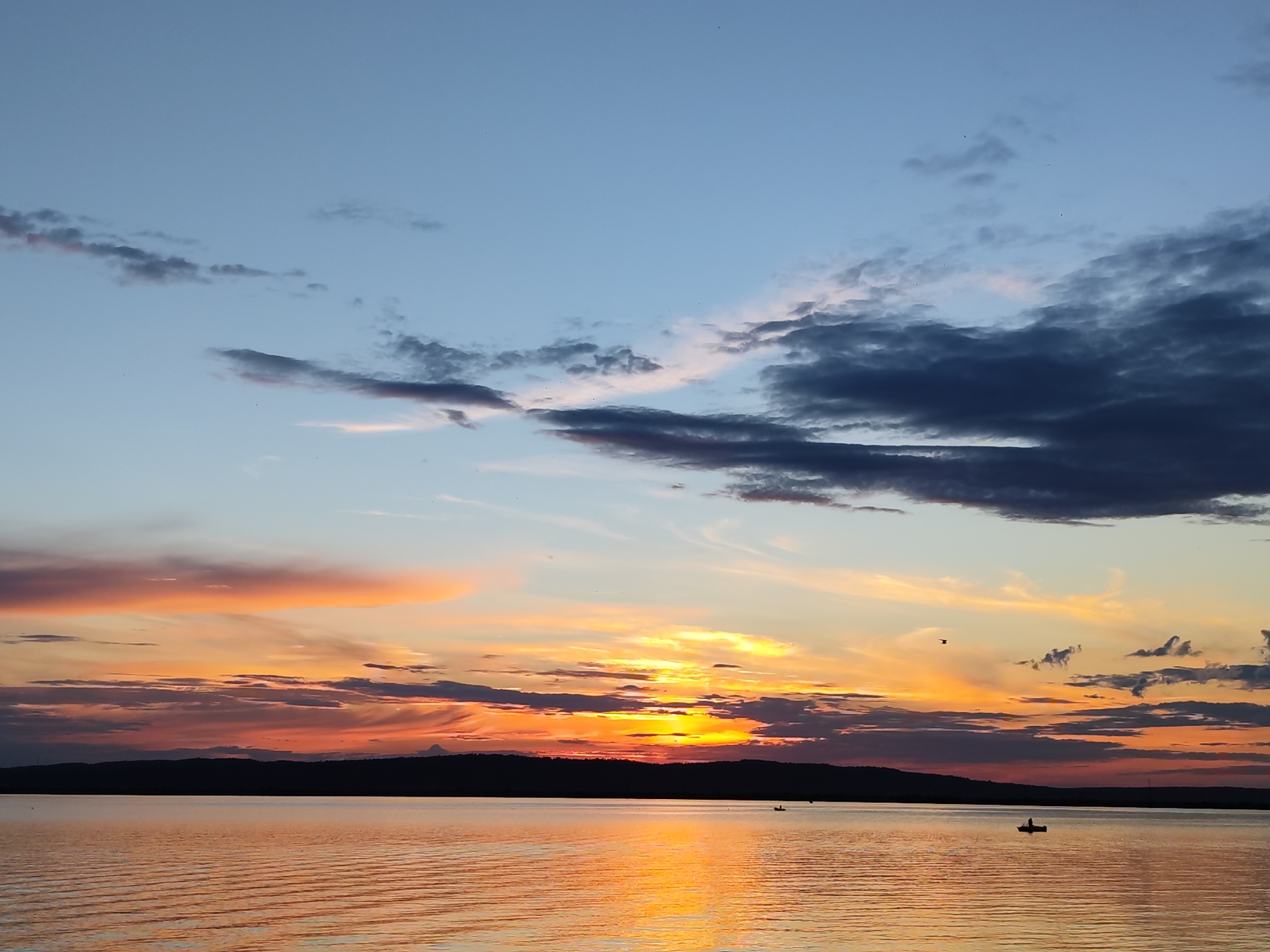 The image depicts a serene sunset over a large body of water. The sky is a canvas of color, transitioning from a soft blue at the top to vibrant oranges and pinks near the horizon. Dark, dramatic clouds are scattered across the sky, particularly on the right side, contrasting with lighter, wispy clouds that catch the sunset's glow.
A dark, silhouetted shoreline, likely a hill or distant landmass, forms a distinct line separating the water from the sky. The water itself reflects the colorful hues of the sunset, creating a shimmering, golden surface with subtle ripples.
Several small objects are visible on the water, appearing to be boats with figures in them, suggesting that people are enjoying the evening on the water. One boat is particularly noticeable in the lower right quadrant, with a lone figure casting a shadow. Another smaller boat is further to the left. A bird is also captured in flight against the colorful sky.
The overall mood of the image is peaceful and tranquil, capturing the beauty of a natural sunset. The time of day is clearly evening, as indicated by the sunset. The presence of a large body of water and a distant shoreline suggests a natural landscape setting, possibly a lake or a wide river. Given the location context, this scene could be the Kama River near Chaikovsky, Russia. No visible text is present in the image.