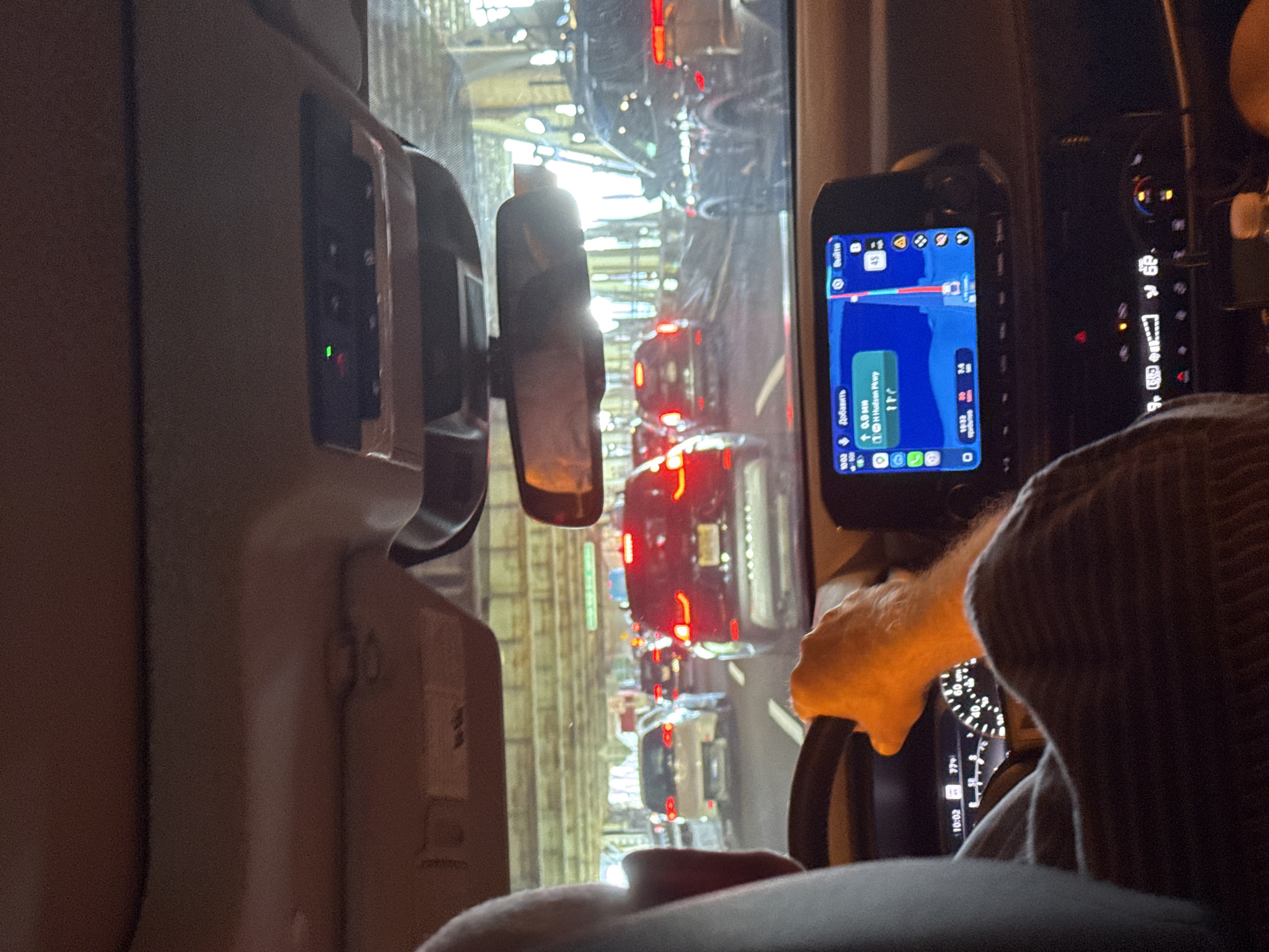 This media file depicts the interior of a vehicle, likely a car, with a view of a traffic jam outside. The scene is oriented vertically, suggesting it was captured with the phone held sideways.
In the foreground, a person's hands are visible, one of which is gripping the steering wheel. The person is wearing a cap. The dashboard is partially visible, featuring a GPS navigation device displaying a route on a map with traffic information. The time on the device reads "10:02" and later "10:32". The text on the GPS indicates "1H Hudson Pkwy", a highway in New Jersey, United States.
Outside the vehicle, a long line of cars is stuck in traffic. The brake lights of the cars are illuminated, indicating a standstill. The environment appears to be urban, with tall structures visible in the background, possibly buildings or overpasses. The lighting suggests it is daytime, but the specific weather conditions are not discernible. The overall mood is one of being stuck in traffic, with a focus on the driver's perspective and the navigation system.