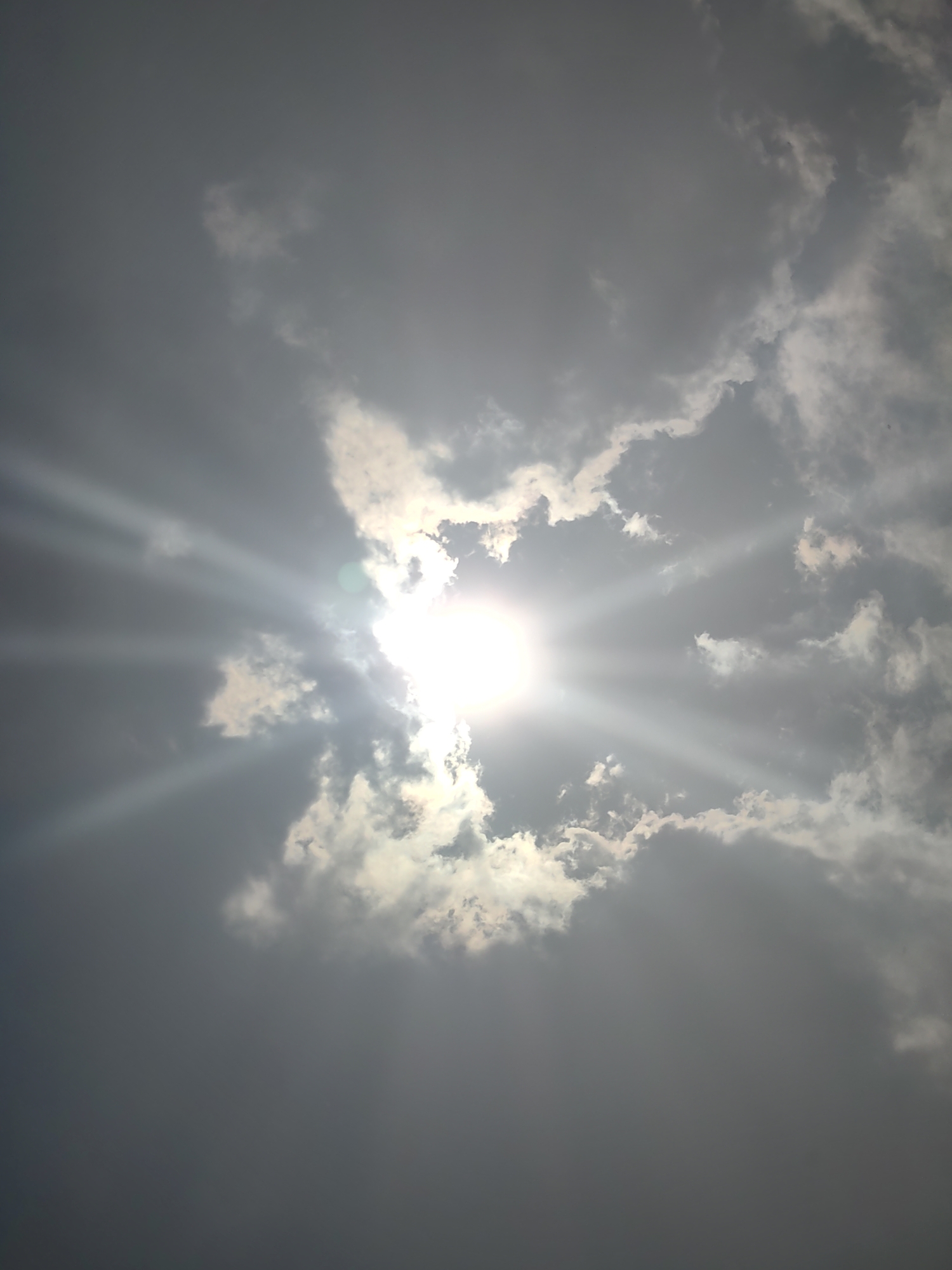 The image shows a sky filled with dark clouds. The sun is peeking through a gap in the clouds, casting bright rays of light. The sun's rays are spread out, creating a starburst effect. The clouds are varied in texture and density, with some appearing thicker and darker, while others are thinner and more wispy, allowing the sunlight to penetrate. The overall impression is one of a dramatic and dynamic sky. This image was captured in Vashisht, India.