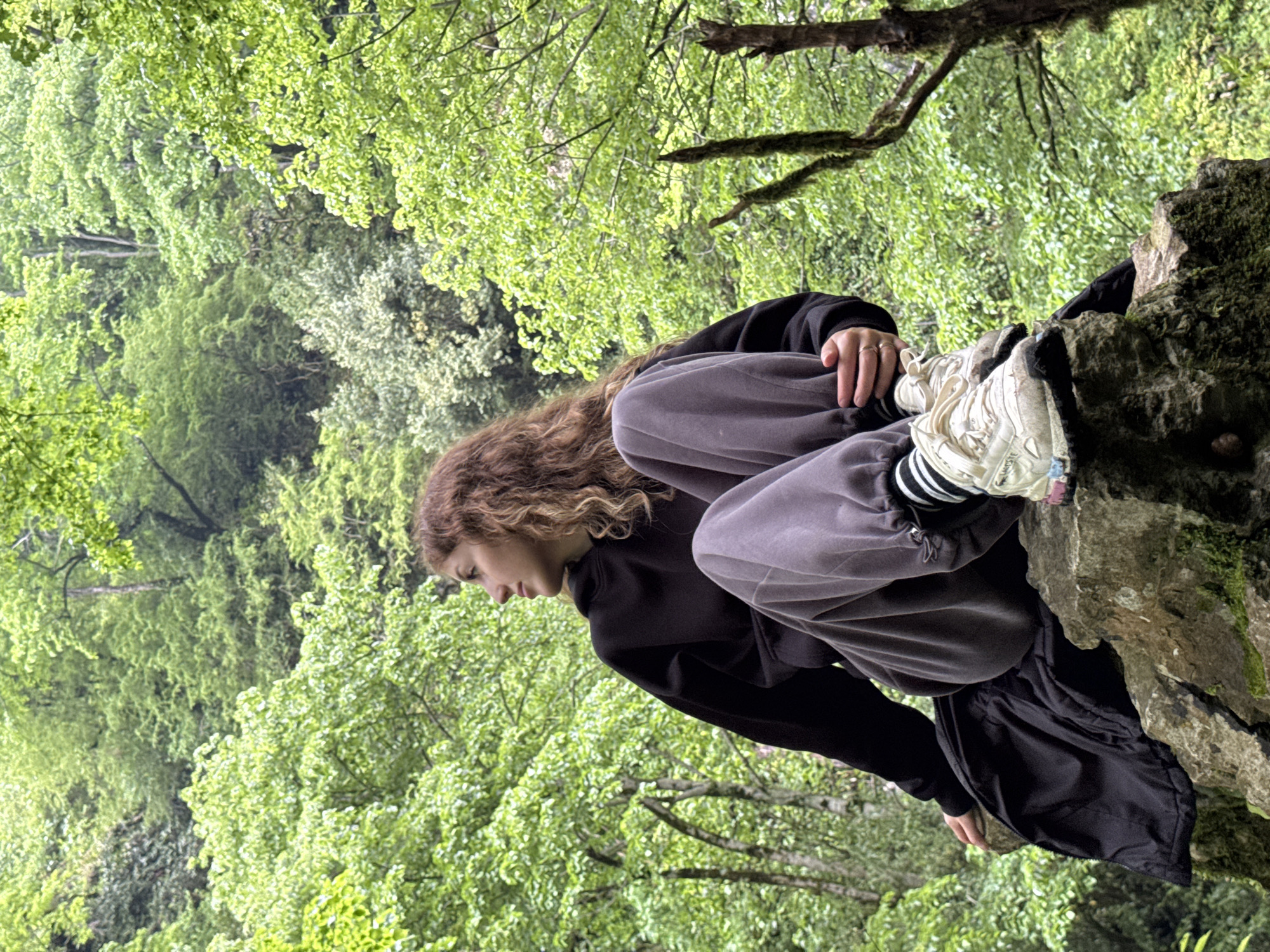 A young person with curly brown hair is perched on a moss-covered rock. They are wearing a black hoodie and gray sweatpants, with white sneakers and black and white striped socks visible. The person is looking down, appearing contemplative or observing something below. The rock is located amidst a lush green forest with trees filling the background. Based on the location information, the scene takes place in Tsabal, Abkhazia.