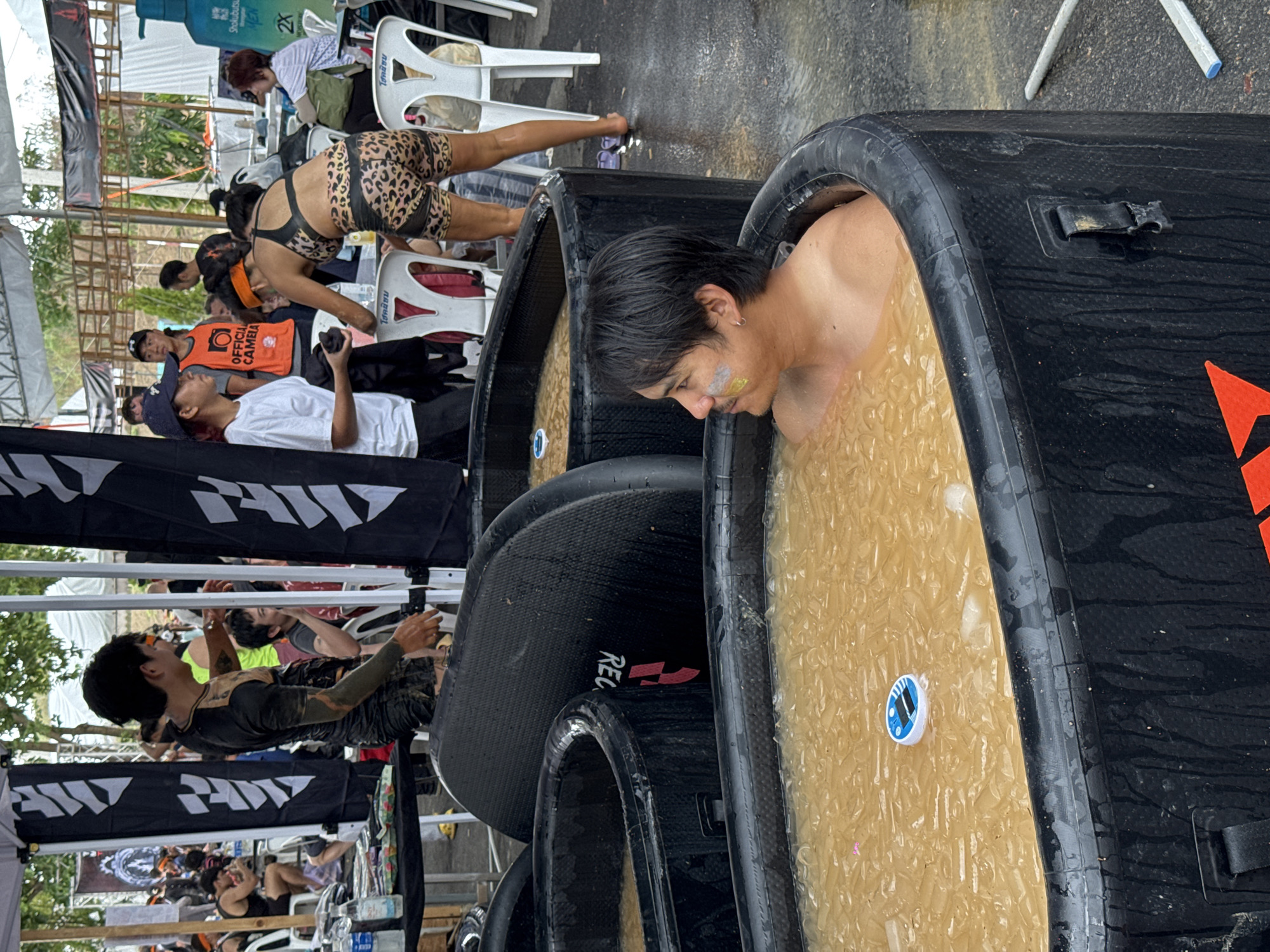 The image depicts an outdoor setting in บ้านสำนักตะแบก, Thailand. Several black inflatable tubs filled with a yellowish liquid and ice are lined up. A man is submerged in one of the tubs, his head and shoulders visible above the waterline. He has dark hair and appears to have some markings on his face. People are milling around in the background, some seated on white plastic chairs. One person is wearing leopard-print shorts and a matching top. A man in a white t-shirt and another man in an orange vest labeled "Official Camera" are also visible. Banners with a white logo on a black background are partially visible in the foreground and background. The environment appears to be a muddy area, likely a recovery zone or similar setup after an athletic event.