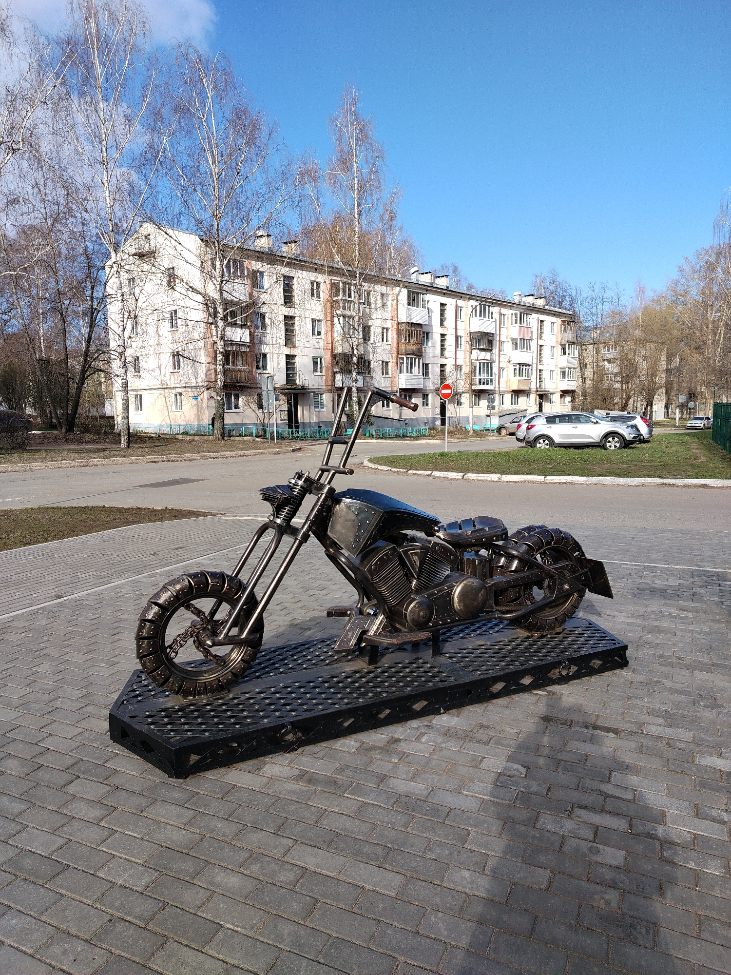 The image shows a metal sculpture of a motorcycle on a paved area, likely a pedestrian zone or plaza, in the city of Chaikovsky, Russia. In the background is a multi-story apartment building, characteristic of Soviet-era urban planning, and some bare trees suggesting it is early spring or late autumn. Several parked cars are visible near the building, and a "no entry" sign stands on the roadside. The motorcycle sculpture sits on a black, metal platform, emphasizing its status as an art installation. The sculpture is highly detailed, with visible elements like the engine, wheels, and seat.