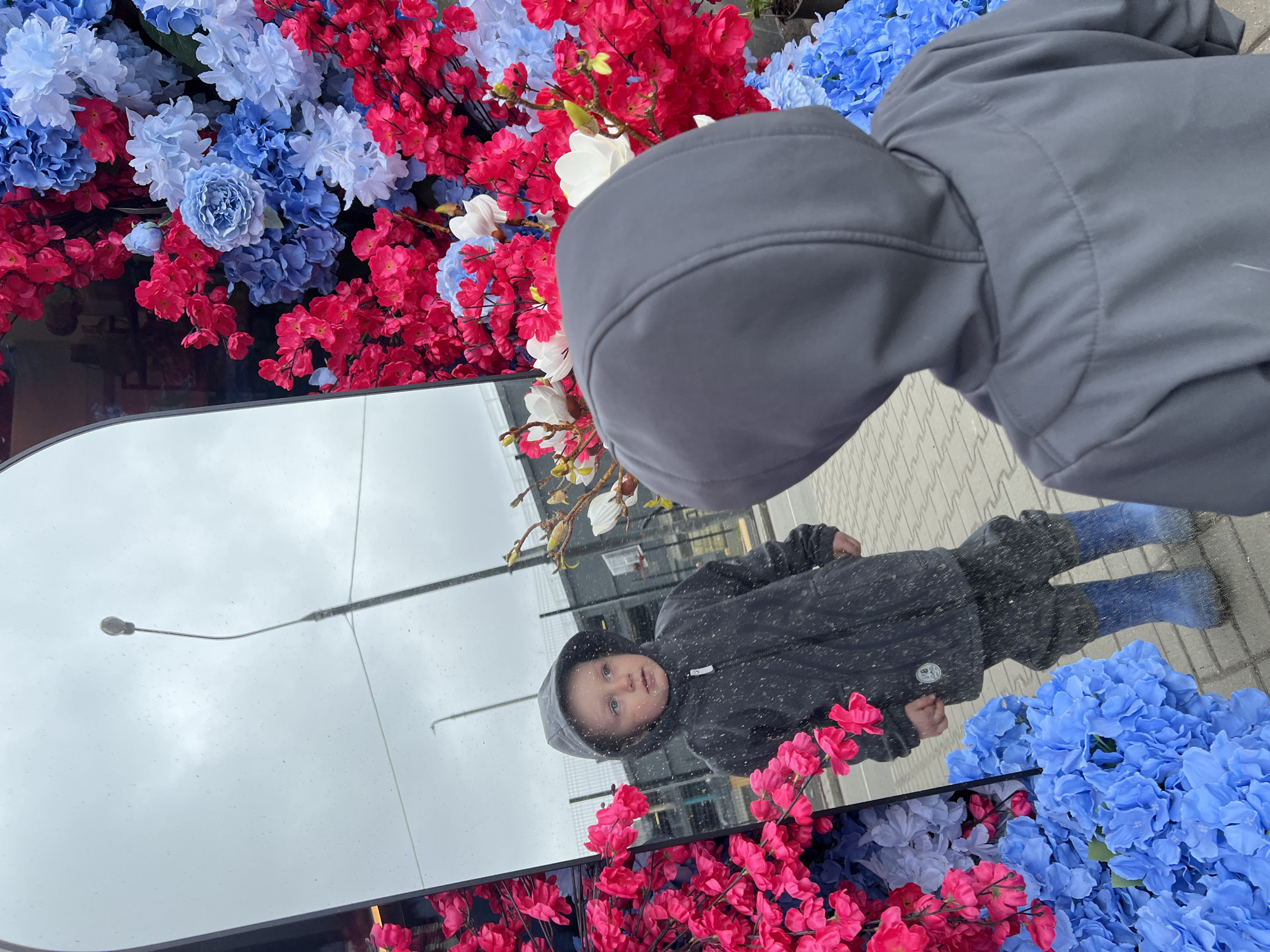 A young child looks into a large, upright mirror, which is bordered by an abundant display of artificial flowers. The flowers are predominantly vibrant red/pink cherry blossoms and various shades of blue hydrangeas, arranged densely around the mirror's frame.
In the foreground, partially visible, is the back of an adult's dark, hooded jacket, suggesting the photographer is with the child. The child, dressed in a dark hooded jacket and blue boots, is clearly reflected in the mirror. Their reflection shows them standing on grey interlocking pavers, looking forward with a curious and calm expression. Small water droplets or dust specks are visible on the mirror's surface.
The mirror's reflection also captures a cloudy, overcast sky and a street light in the upper portion, indicating a daytime setting. The scene takes place outdoors in a public area, possibly a decorative installation or storefront display, located in Mytishchi, Russia. There is no discernible text visible in the image.