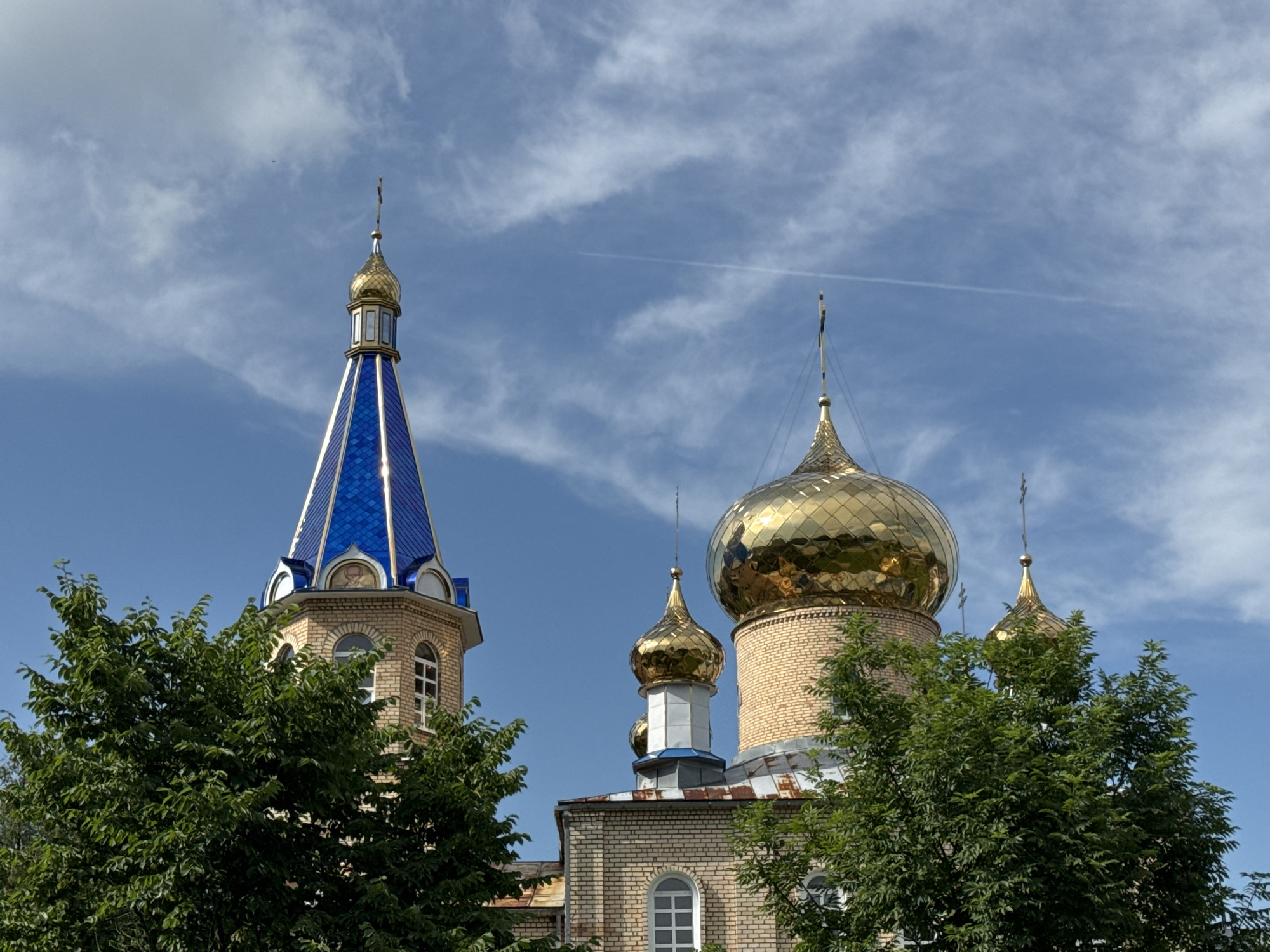 The media file captures an exterior view of a church in Cherven, Belarus, on a clear day. The main subjects are the church's towers and golden domes, set against a bright blue sky dotted with wispy white clouds. The architecture features light-colored brickwork, with a tall, slender tower topped with a blue, faceted roof and a golden spire. Adjacent to this, several rounded golden domes rise, some larger and more prominent than others, each crowned with a cross. Green trees with dense foliage partially obscure the lower sections of the church building and frame the scene. The sunlight suggests it is daytime, likely late morning or afternoon, given the brightness and the clear sky. There is a faint contrail from an airplane visible in the sky. No people or specific events are depicted. The image focuses on the serene and majestic appearance of the religious structure.