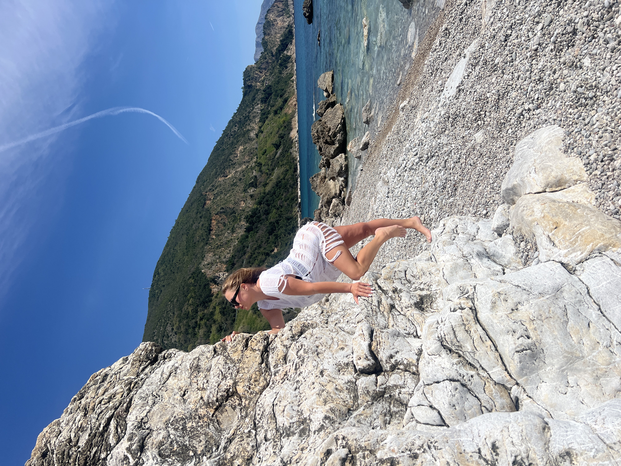 A medium shot shows a woman doing a push-up on a light gray rock formation. The woman is wearing a white, fringed dress and sunglasses. She is on a beach with small, gray pebbles, turquoise water, and a mountain covered in green trees. The sky is blue with a white contrail in the upper left corner. The location is in the city of Prijevor, Montenegro.