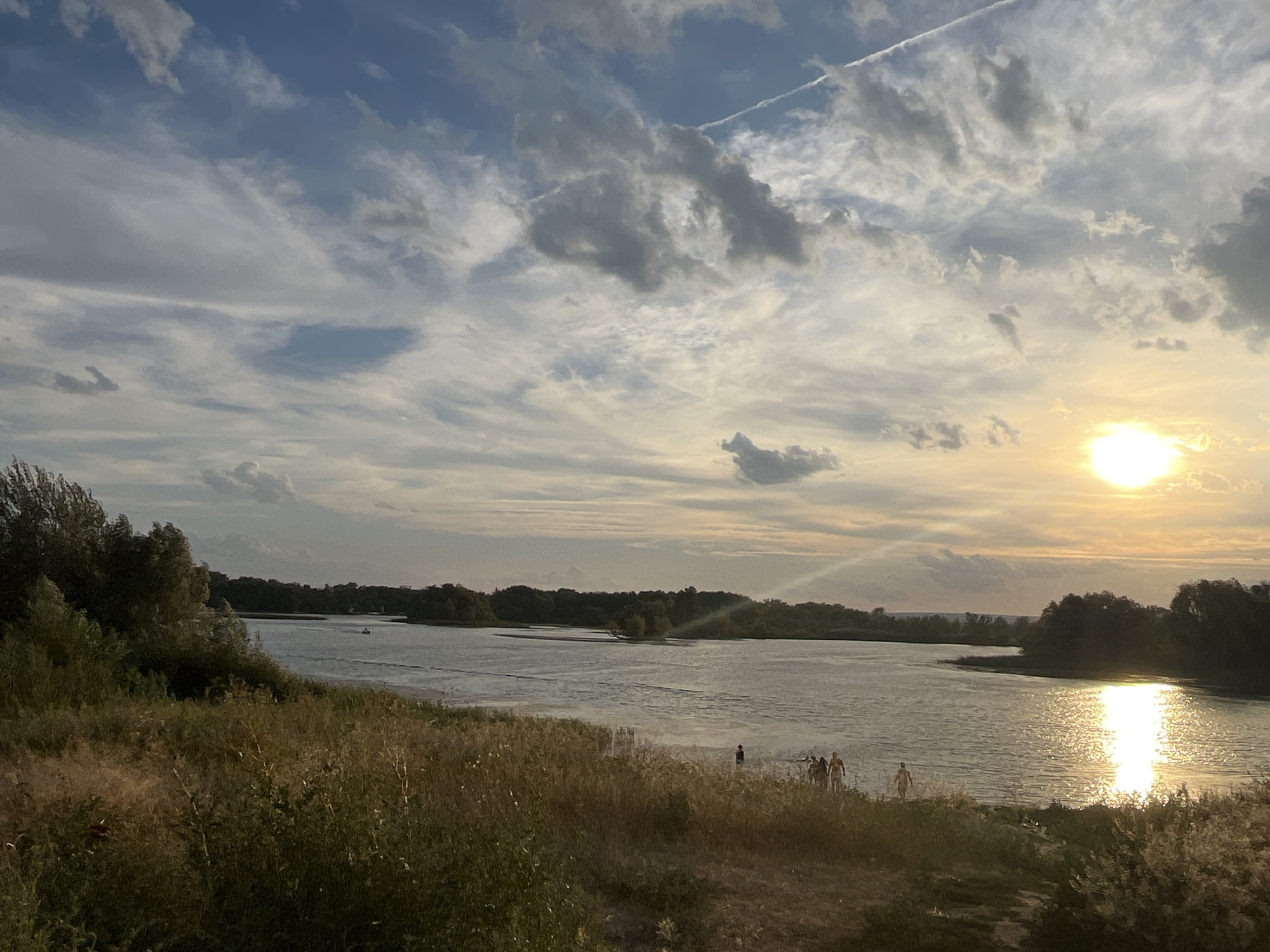 A wide shot captures a serene natural landscape at sunset, likely near the village of Shumeyka, Russia. The scene is dominated by a large body of water, reflecting the warm, golden light of the setting sun. The sky above is a dramatic canvas of blue, with scattered clouds in various shades of grey and white, some illuminated by the sun's rays. A thin, white contrail is visible streaking across the upper portion of the sky.
The foreground is characterized by a grassy embankment, with dry, tall grasses and some green foliage. On the shoreline, a small group of people, appearing to be four individuals, are standing near the water's edge. One person is standing slightly apart from the others. The water itself appears calm, with ripples indicating slight movement. Across the water, a distant treeline marks the opposite bank, densely covered with lush green vegetation. A small boat is visible on the water in the mid-ground, moving away from the viewer. The overall atmosphere is peaceful and tranquil, suggesting an end-of-day scene.
