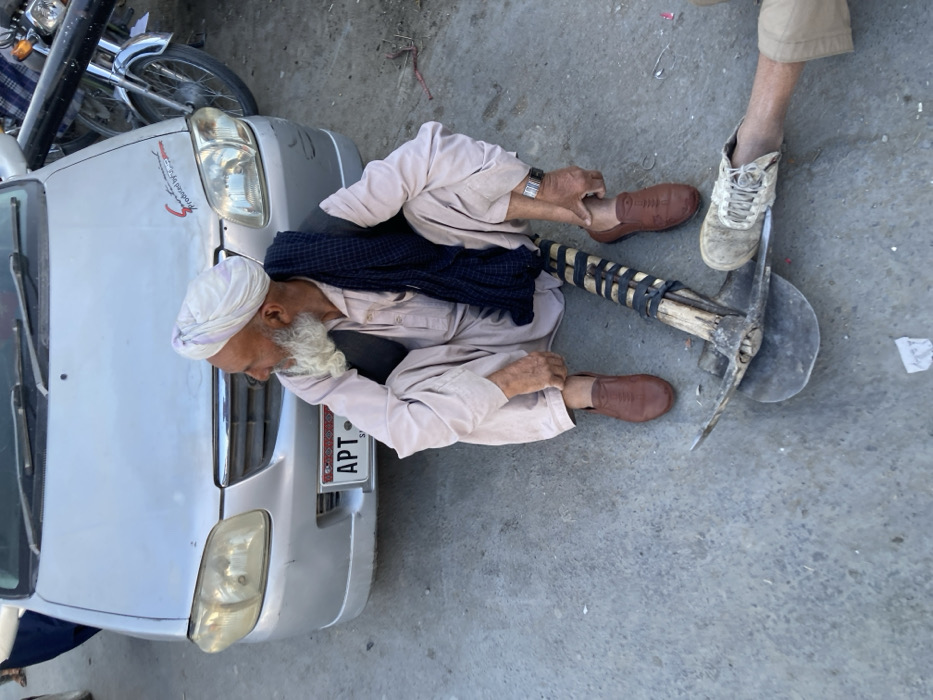 The image depicts an outdoor scene in Quetta City Tehsil, Pakistan, showing an elderly male seated on a concrete surface next to a parked silver car.
The primary subject is an elderly male with a long white beard and a white turban. He is dressed in a light beige shalwar kameez, a dark vest, and a dark blue shawl draped over his left shoulder. He wears brown slip-on shoes and a silver-colored watch on his left wrist. He is seated with his knees bent, looking towards the left side of the frame with a slight smile. His left hand is positioned near his left ankle.
Behind the man is the front of a silver-colored car, appearing to be a Suzuki Alto. The license plate, partially visible, reads "APT" and "SIN" below, indicating a Sindh province registration. A red sticker with "Sport minded" is affixed to the front bumper. To the far right, part of a motorcycle, including a chrome fender and front wheel, is visible.
Between the seated man and a partial view of another person's lower leg and foot, a digging tool resembling a pickaxe with a shovel blade is resting on the ground. The tool has a wooden handle reinforced with dark strapping. The partial foot, wearing an off-white lace-up shoe and khaki-colored pants, is positioned over the blade of this tool.
The ground is concrete, showing minor debris including small pieces of white paper and reddish twigs. The scene is illuminated by natural light, casting subtle shadows.
