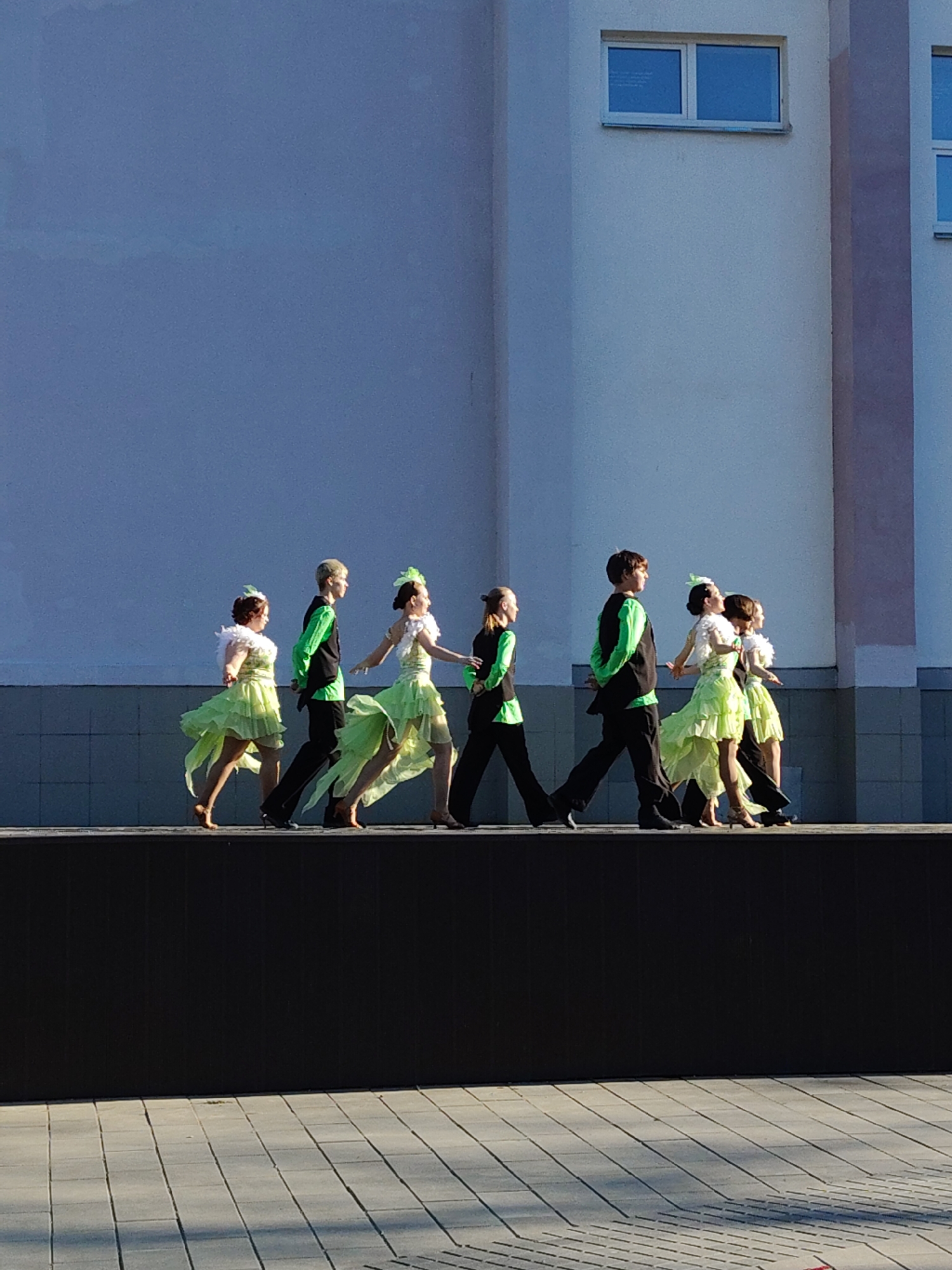 In an outdoor setting in an unidentified city, a group of seven young dancers are seen walking in a single line on a dark-colored platform, which is likely a raised stage or a section of a public space. The dancers consist of both male and female individuals. The female dancers are wearing light green dresses, some of which appear to be high-low style. The male dancers are wearing black pants and vests, and bright green shirts. They are all walking at the same pace, with the dancers' movement appearing synchronized. Behind the dancers is a light-colored building with two windows visible on its upper portion. The ground is paved with rectangular concrete blocks. The scene appears to be set in daylight, with shadows cast on the platform and the paving, suggesting a sunny day.