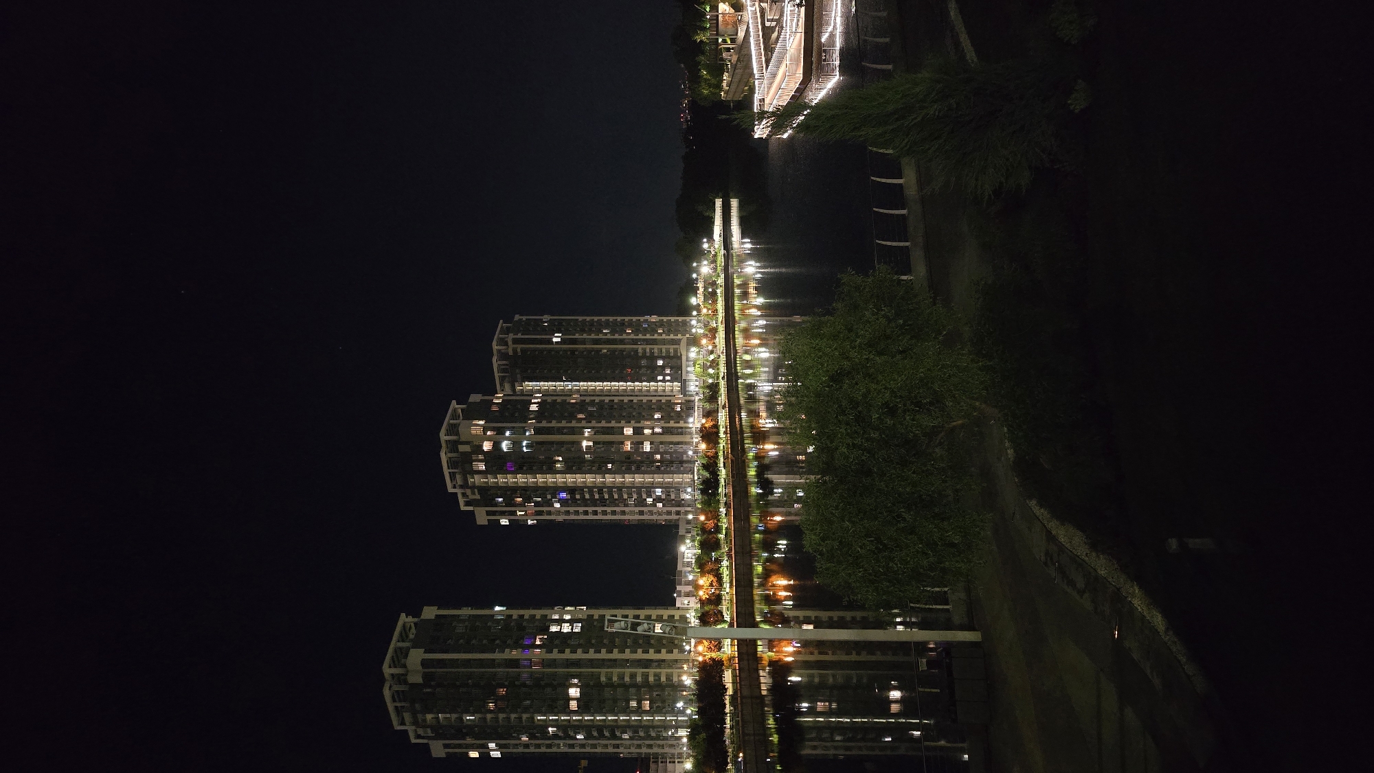 The image presents a tranquil night scene in Kyiv, Ukraine, focusing on an urban waterscape. Tall, modern apartment buildings dominate the right side of the frame, their numerous windows illuminated with various lights, suggesting active dwellings. The architecture features distinct, multi-level designs.
A brightly lit promenade or pathway runs along the base of these buildings, separating them from a calm body of water—likely a river or canal. This walkway is lined with a series of evenly spaced streetlights, which cast strong, elongated reflections onto the dark surface of the water. The reflections of the building windows and the promenade lights create a captivating shimmer across the water, mirroring the urban glow above.
On the opposite bank of the water, to the left, dark masses of vegetation, including trees and bushes, are visible. These natural elements offer a contrast to the artificial brilliance of the city lights. The sky above is a deep, clear black, characteristic of a night scene.
There are no people or specific activities depicted, lending a sense of stillness and peacefulness to the urban environment. The primary emphasis is on the interplay of light and reflection, highlighting the serene beauty of the city after dark. No visible text is present in the image.