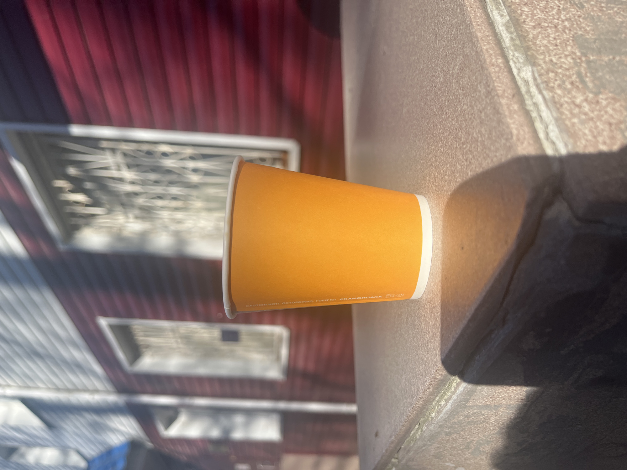 An eye-level, horizontally-oriented, sun-lit image depicts an orange paper coffee cup with a white rim resting on a concrete surface. Behind the cup, a blurred red, white, and gray exterior of a building is visible, along with the suggestion of a lattice-patterned window. The base of the cup is closer to the viewer than the rim. The location is the city of Ulyanovsk, Russia. There is no indication of people in the scene. The cup has warning text printed on its exterior in both English and Russian: "CAUTION HOT! OСТОРОЖНО! ГОРЯЧО! СКАНДИНПАКК". The cup rests on a concrete wall with shadows.