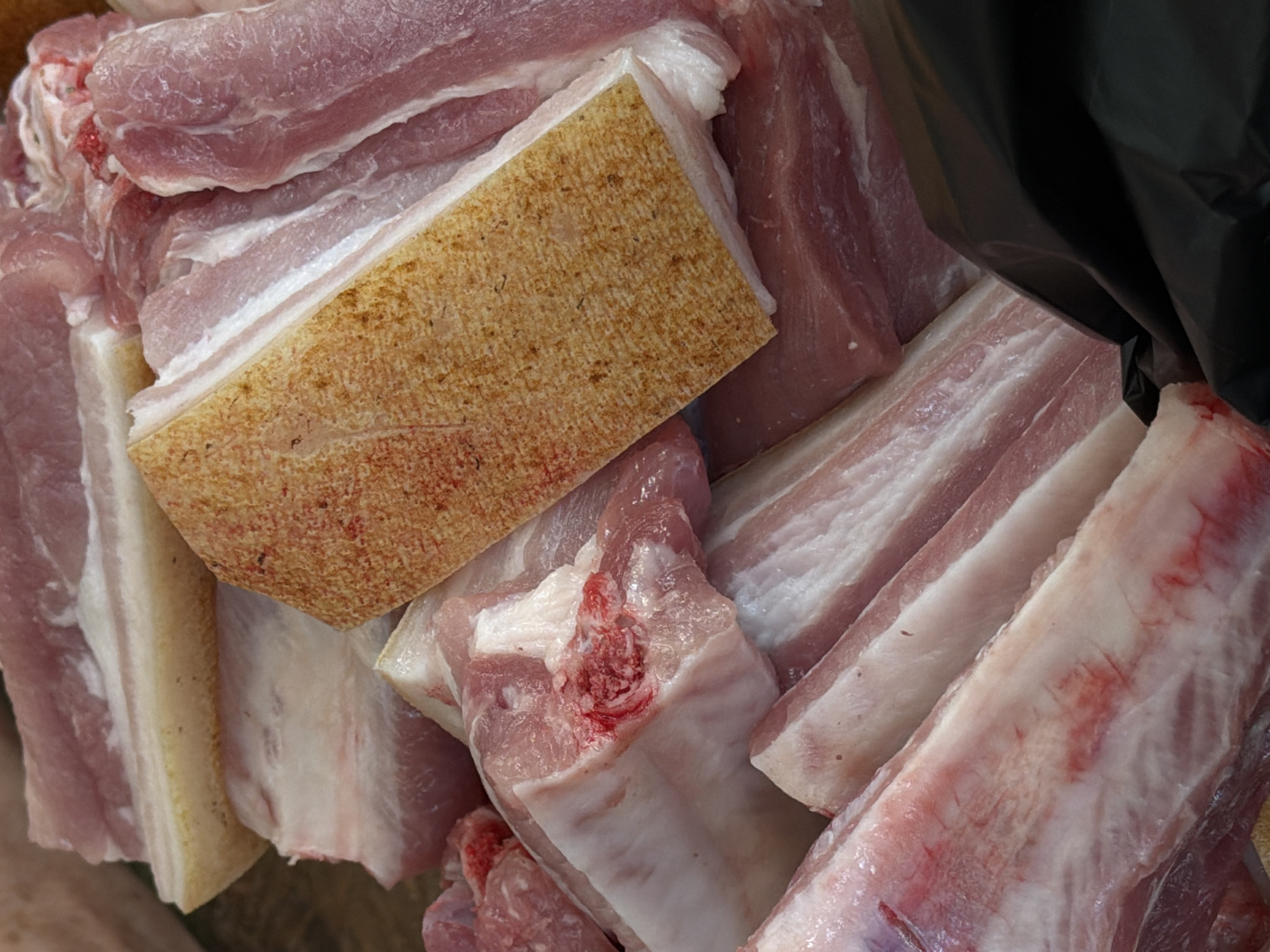 The image shows a close-up, overhead view of a pile of raw pork cuts, some with skin on. The meat is a pale pink color with streaks of white fat. The skin is a light tan color with a slightly rough texture. Some of the cuts are large and rectangular, while others are smaller and irregularly shaped. The meat appears to be fresh and has been recently butchered. The background is dark and out of focus, with a hint of a black plastic bag visible on the right side. The scene is likely from a butcher shop or a market stall. No specific location in Tuapse, Russia can be identified from the image alone.