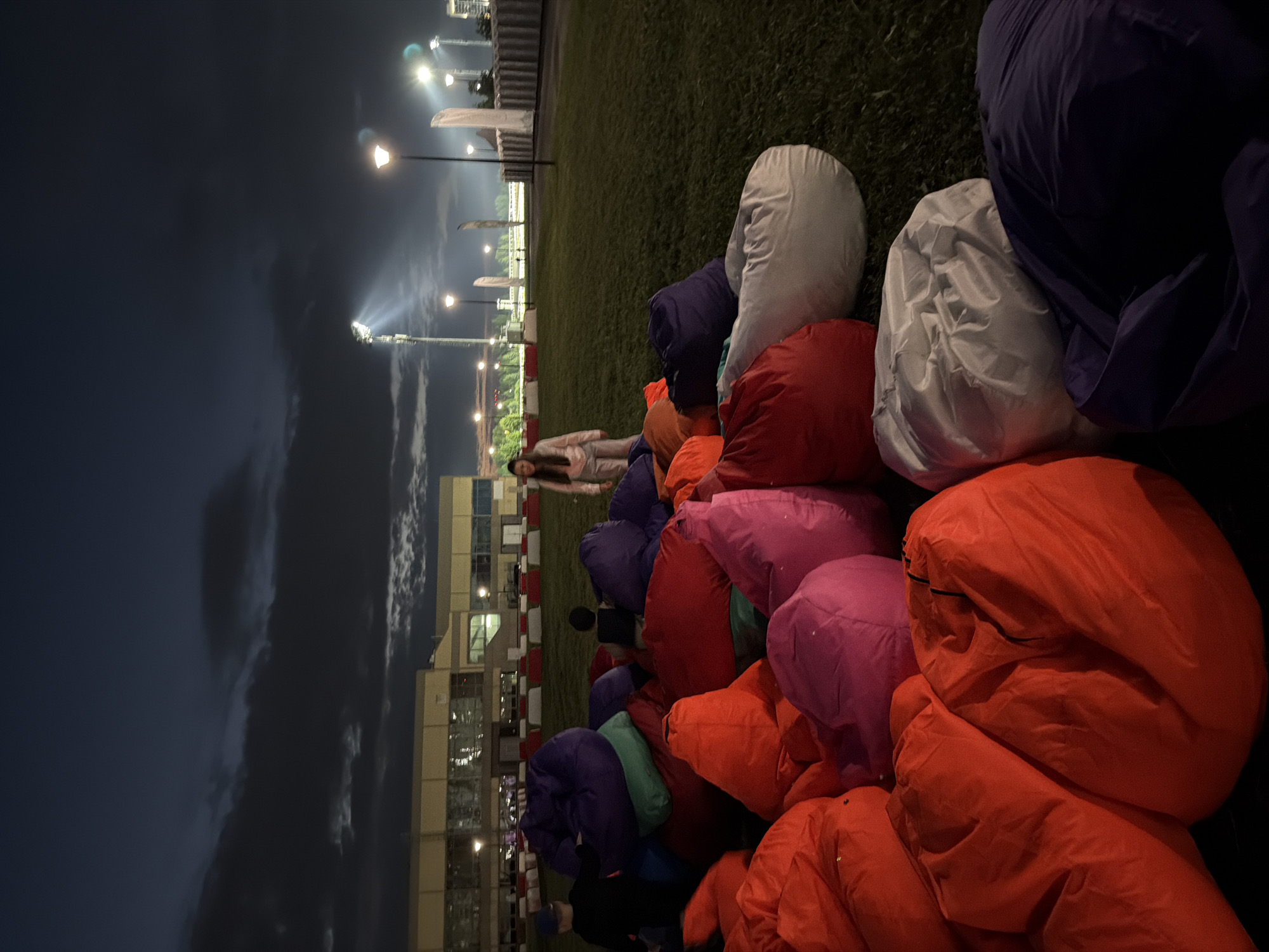 An outdoor scene at dusk or night with a field of colorful beanbag chairs on a grassy area. In the background, a large building with many windows is illuminated, and stadium lights are visible, casting bright beams into the dark sky. Several people are present, including a young woman standing in the middle of the beanbag chairs and two children, one wearing a blue cap, interacting with the chairs. The beanbag chairs are in various colors including red, orange, pink, purple, green, and white. The overall atmosphere suggests an outdoor event or gathering.