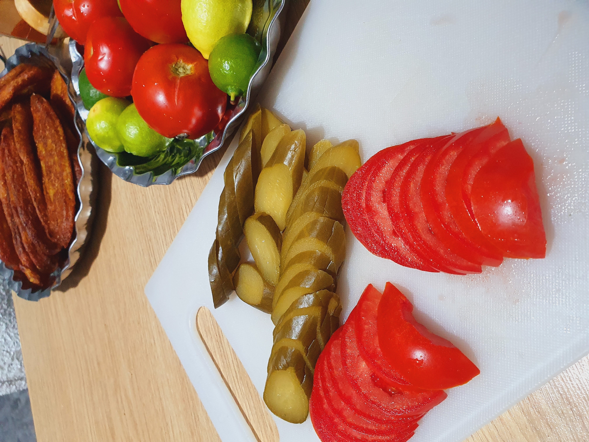 In Kheyrabad, Iran, a close-up, eye-level shot showcases a food preparation scene. The focus is on a white cutting board where sliced tomatoes and pickles are arranged. In the background, a silver bowl holds red tomatoes and green limes, while another metallic bowl contains fried food, possibly churros, with a textured surface. The subjects are purely edible items, and no people or interactions are visible. The environment appears to be a kitchen or dining area, with a wooden table surface visible. The lighting is natural, highlighting the textures and colors of the food.