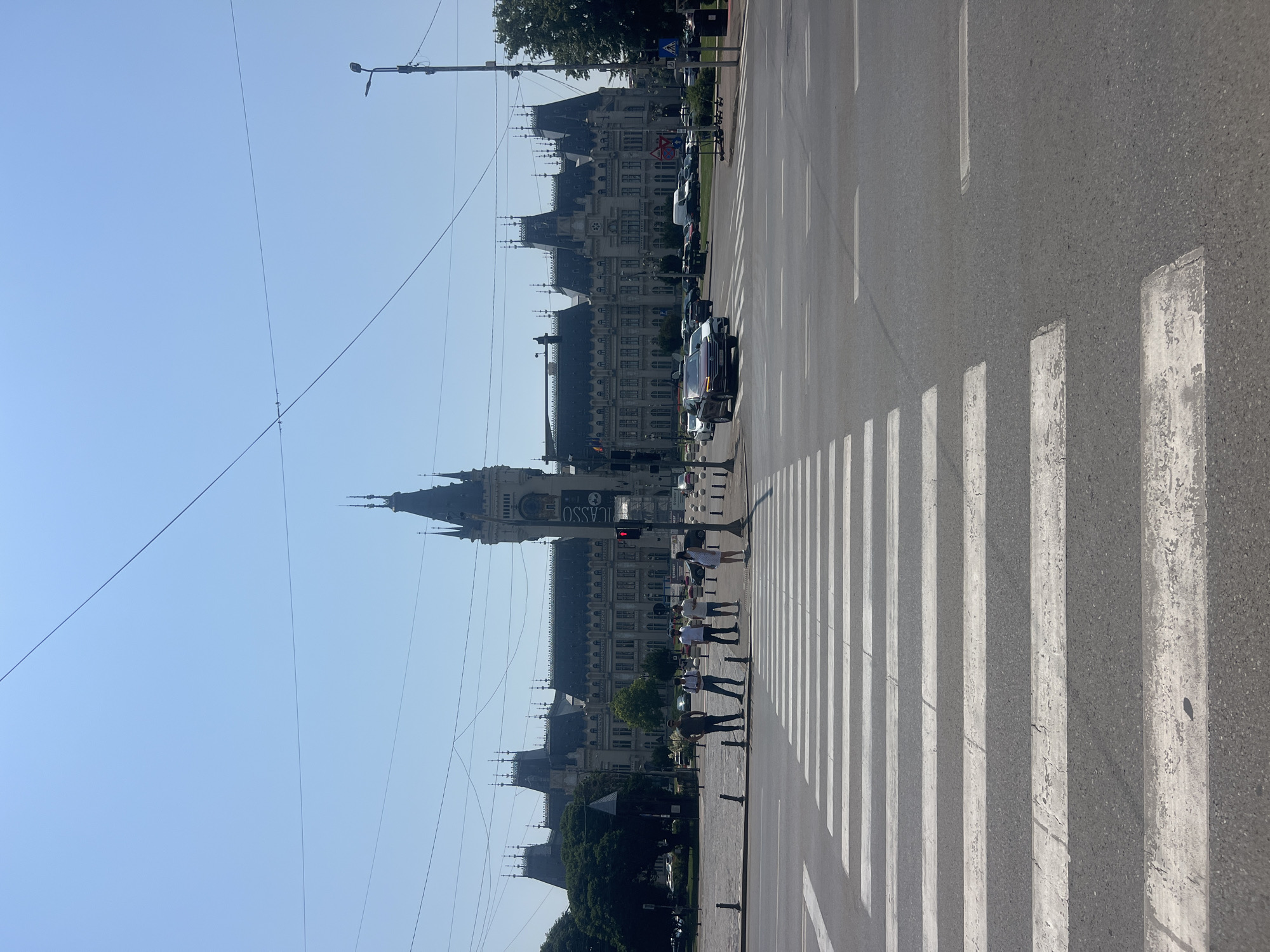 The image shows a wide street intersection in front of the Palace of Culture in Iași, Romania. A pedestrian crossing with white stripes is prominent in the foreground. Several people are standing on the sidewalk near the crossing, appearing to wait or observe. Behind them, a large, ornate building with multiple towers and spires, identified as the Palace of Culture, dominates the background. The sky is clear and blue. Traffic, including cars and vans, is visible on the street. Streetlights and overhead wires are also present, indicating an urban environment. The overall scene depicts a sunny day in a city center, with the grand architectural landmark as the focal point.