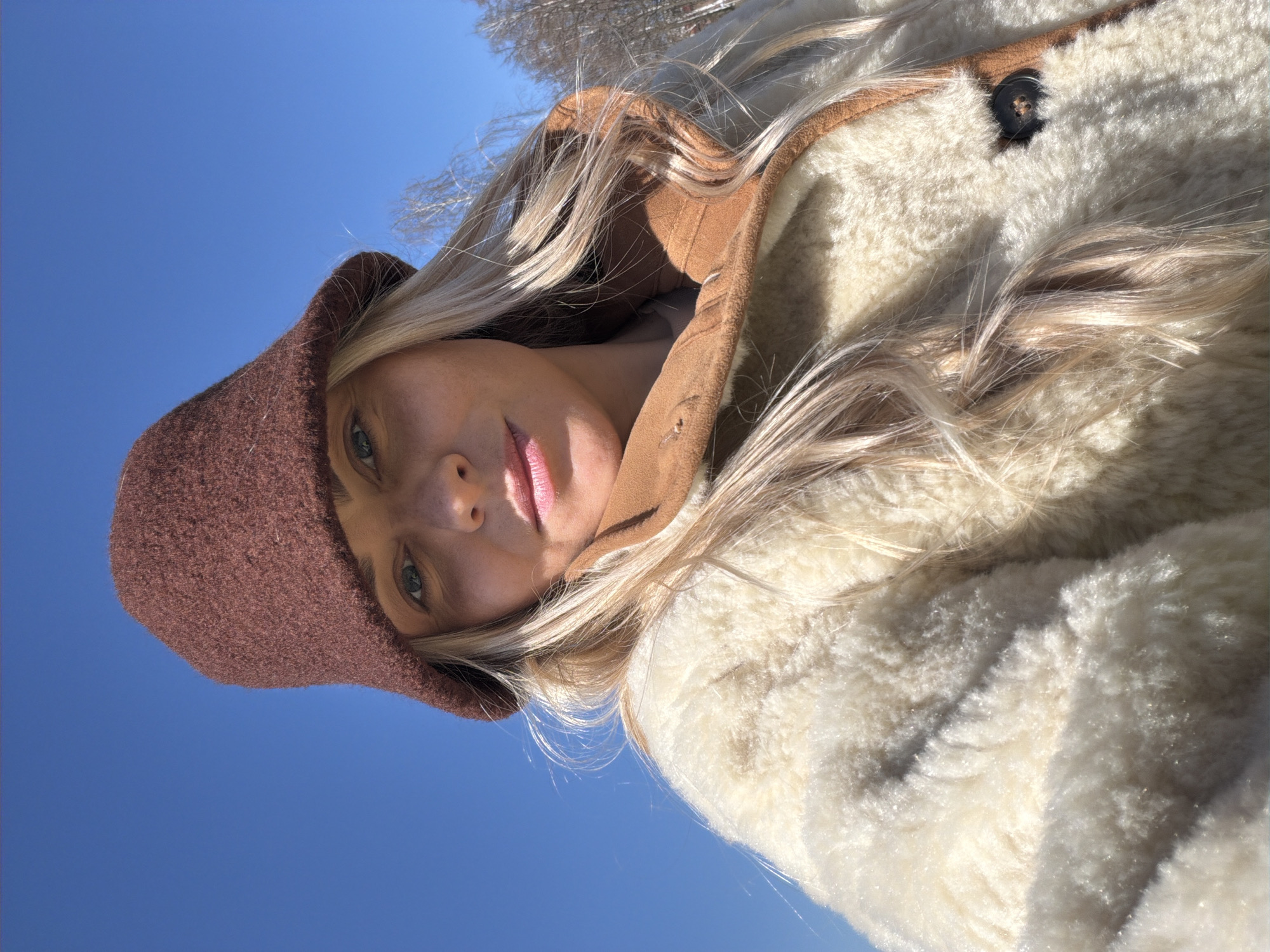 A woman is captured in a close-up shot against a bright blue sky. She is wearing a brown knit hat and a light-colored, possibly sheepskin, coat with brown trim. Her blonde hair is visible beneath the hat and around her face. The perspective is angled upward, with only a small portion of what appears to be tree branches in the upper portion of the frame. The location is unknown.