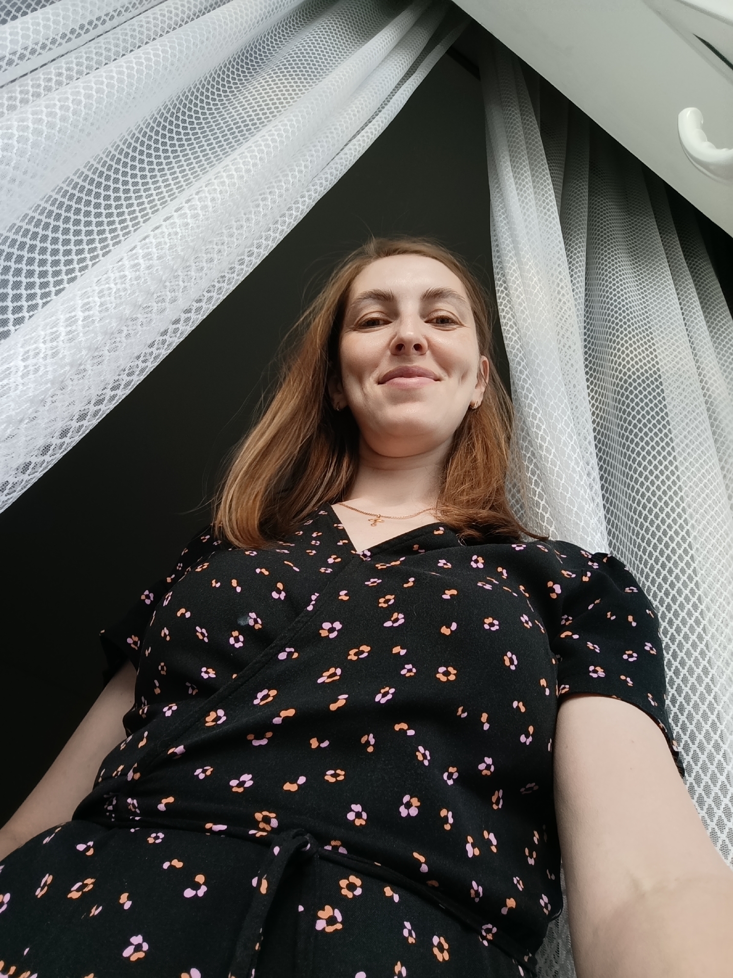 A woman with light brown hair and a dimpled smile is the subject of the photograph. She is wearing a black wrap dress adorned with a pattern of small, stylized flowers in shades of pink and orange. A delicate gold necklace with a small pendant is visible around her neck. She appears to be standing indoors near a window draped with white, net-like curtains. A partially visible white window frame can be seen in the upper right corner. The background is dark, suggesting an interior space with subdued lighting. Based on the user's prompt, the location is identified as Chaikovsky, Russia.