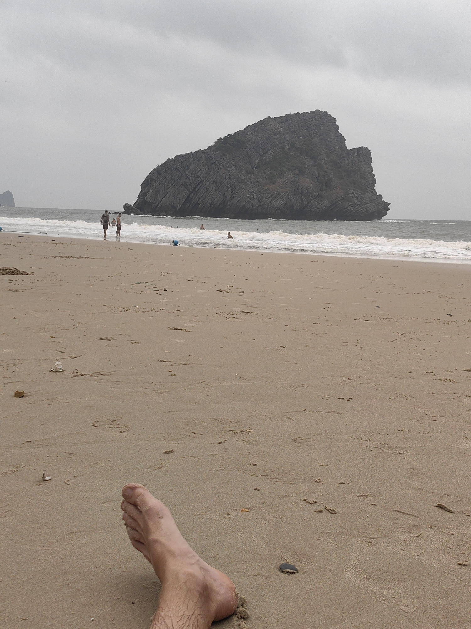 The scene depicts a beach in Hải Phòng, Vietnam, characterized by gray skies and a rocky island in the distance. The foreground is dominated by sand, upon which lies a human foot, likely belonging to the photographer. Further up the beach, there are at least 2 people walking along the shore. In the background, waves are visible, as well as a large dark, rocky island. The overall atmosphere of the scene appears to be overcast and tranquil.