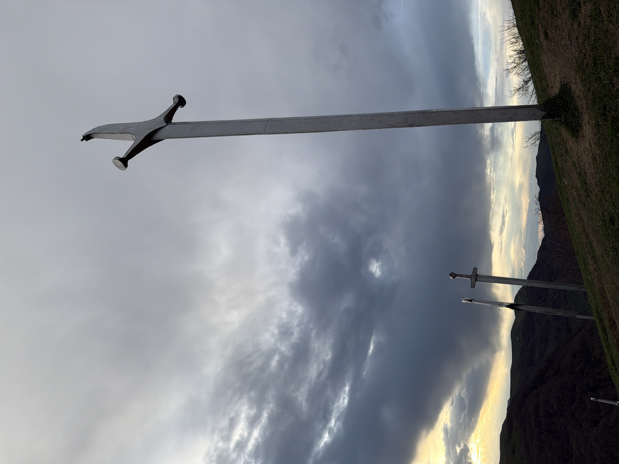 The image shows a landscape with a focus on several monumental metal swords erected vertically into the ground. The swords have a stylized appearance, with large, simplified blades and crossguards. The sky is overcast with dramatic, swirling clouds, ranging from light gray to dark gray hues. In the background, a hillside or mountain is visible, partially covered in vegetation. The location is the city of Zemo Nichbisi, Georgia.