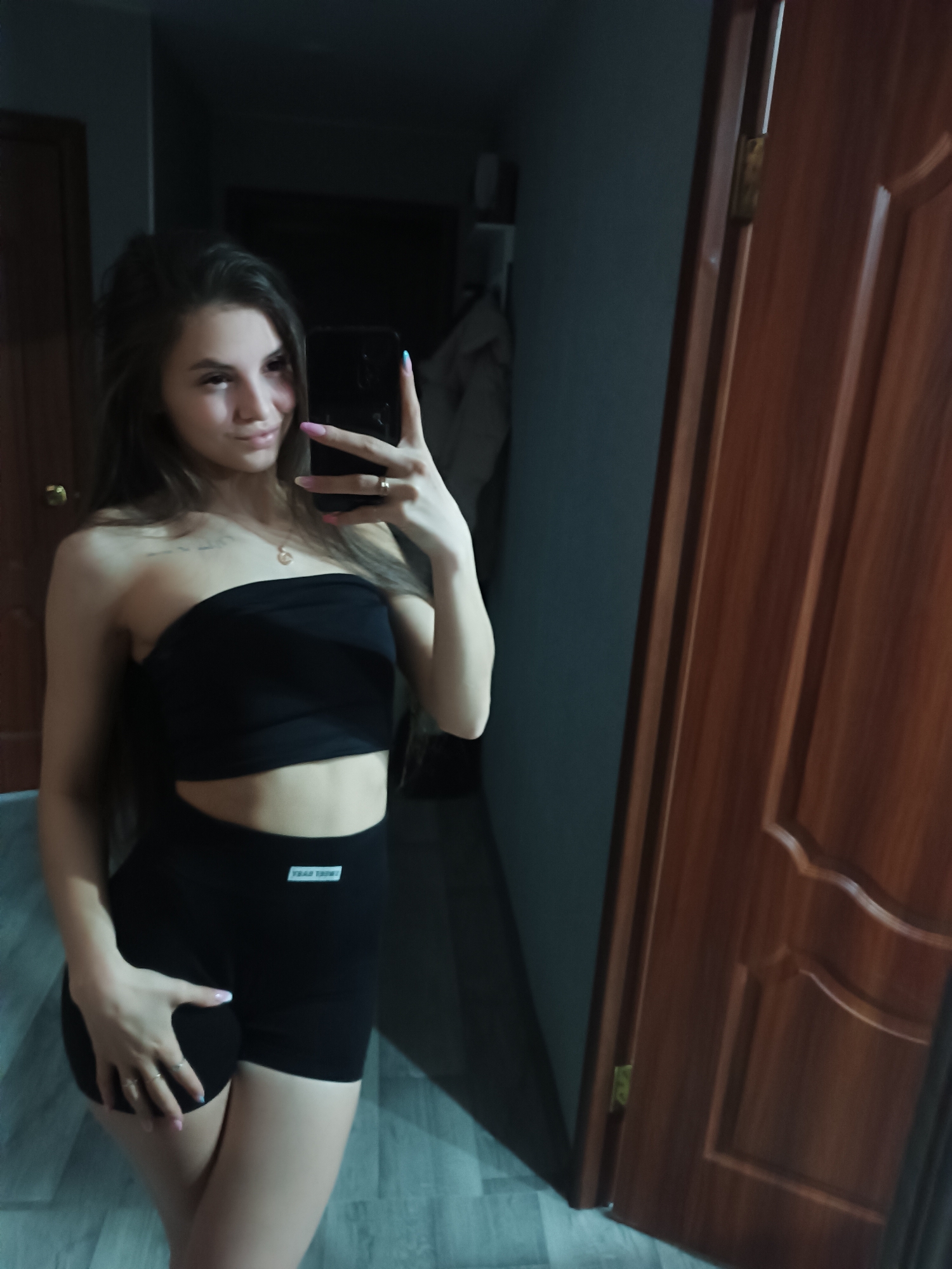 A young woman with long brown hair is taking a selfie in what appears to be an apartment. She is wearing a black strapless top and black shorts. Her left hand is on her hip. In the background are two wooden doors, one partially open, revealing a dimly lit room with a coat hanging. The location appears to be a residential interior, possibly in the city of Chaikovsky, Russia. The floor has a gray wood pattern.