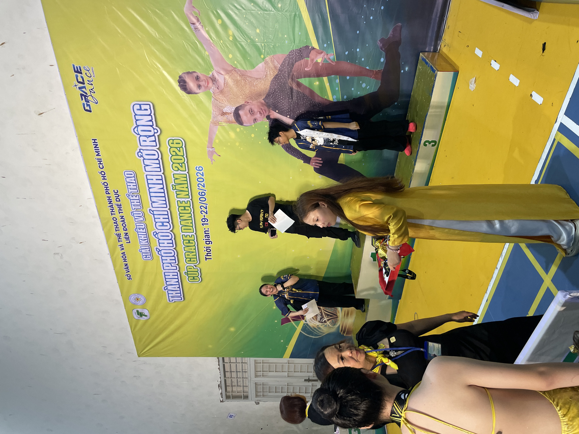The image depicts an awards ceremony or competition taking place in Thuận An city, Vietnam. A large yellow banner serves as the backdrop, featuring text in Vietnamese and the logo "GRACE Dance Academy." The banner prominently displays the title "GIẢI KHIÊU VŨ THỂ THAO THÀNH PHỐ HỒ CHÍ MINH MỞ RỘNG - CÚP GRACE DANCE NĂM 2026" and the dates "19-22/06/2026."
In the foreground, several individuals are present. On the right, a person in a bright yellow áo dài is presenting a tray with trophies to a young woman. Another person, dressed in black with a yellow scarf, stands to the left of the presenter, holding a document. Behind them, a woman in a blue and gold jacket is laughing and holding papers. Further to the left, two young women are conversing, with one facing away from the camera and wearing a yellow garment. A young man wearing a black t-shirt and glasses stands on a podium labeled with the number "3" to the right of the banner. He holds a piece of paper. A large printed image of a dancing couple is visible on the banner behind the participants. The environment appears to be an indoor venue with a yellow floor marked with white lines.