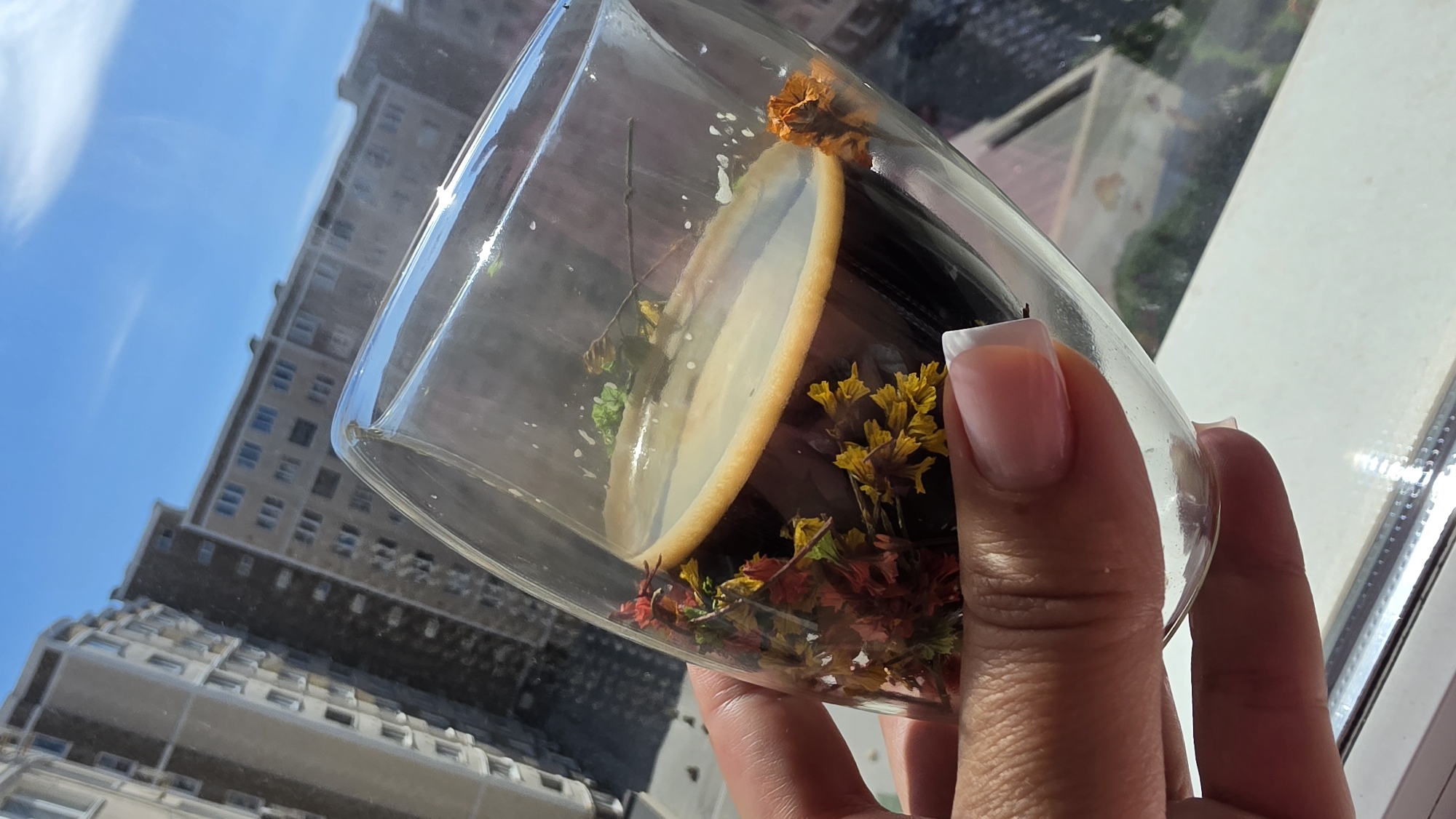 A hand holds a double-walled glass filled with dark coffee and a thin layer of crema. Dried flowers and foliage in red, yellow, and green are visible within the glass, between the coffee and the outer wall. The background features the facade of apartment buildings against a clear blue sky, suggesting an urban setting. The image is a close-up shot, focusing on the beverage and the hand holding it.